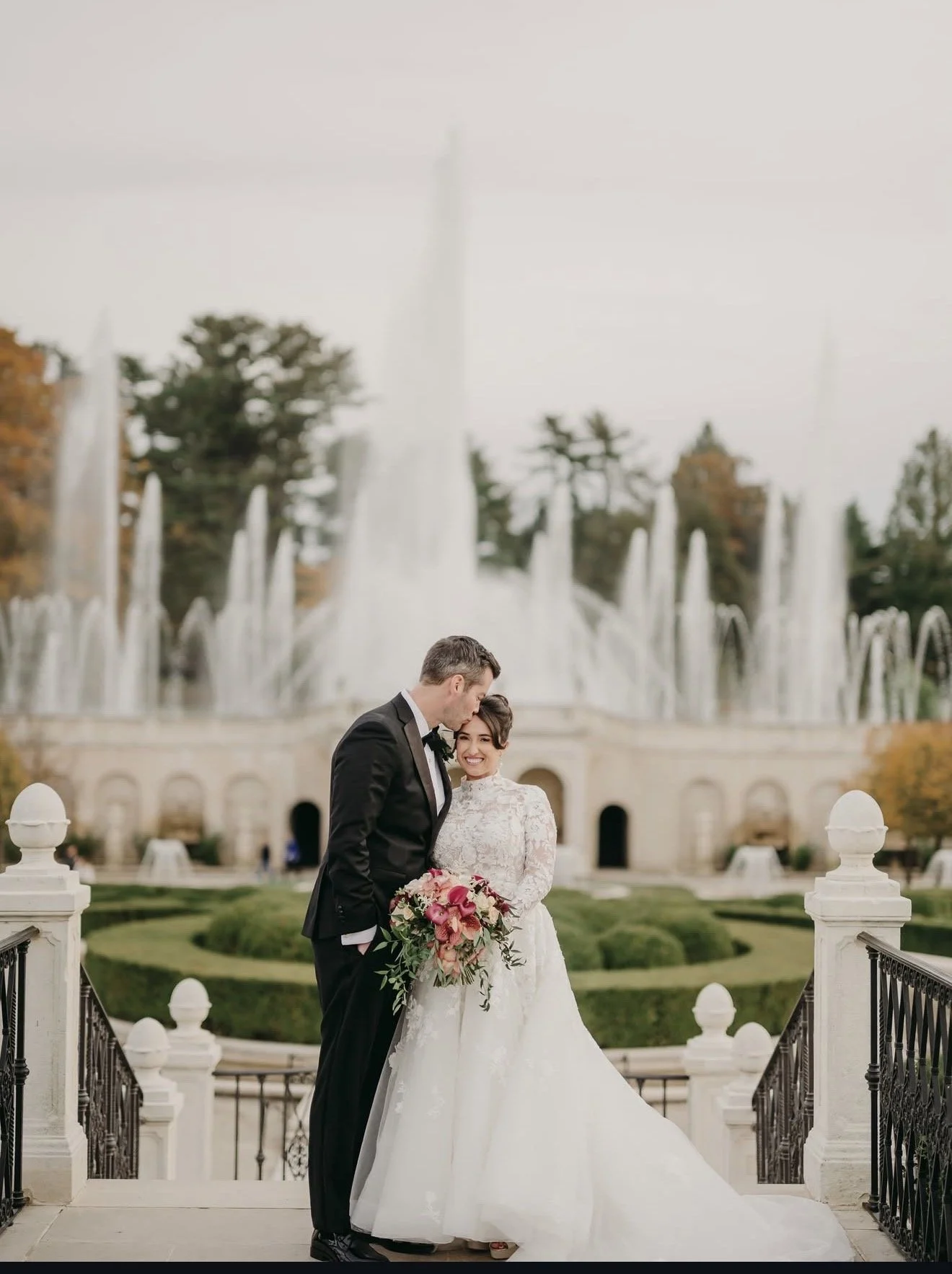 A bride and groom standing together outdoors near a fountain, with the groom kissing the bride on her forehead, during a wedding photoshoot.