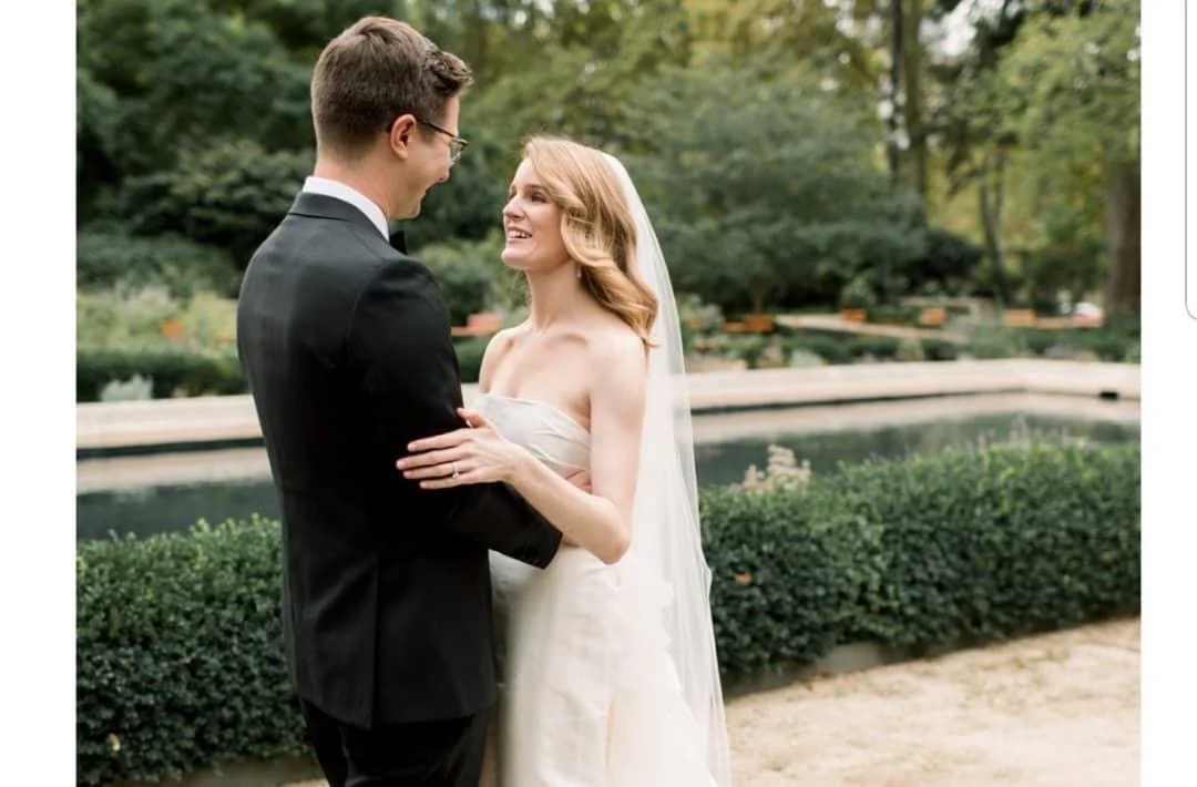 A bride and groom happily exchanging vows outdoors near a pond, surrounded by greenery and trees.