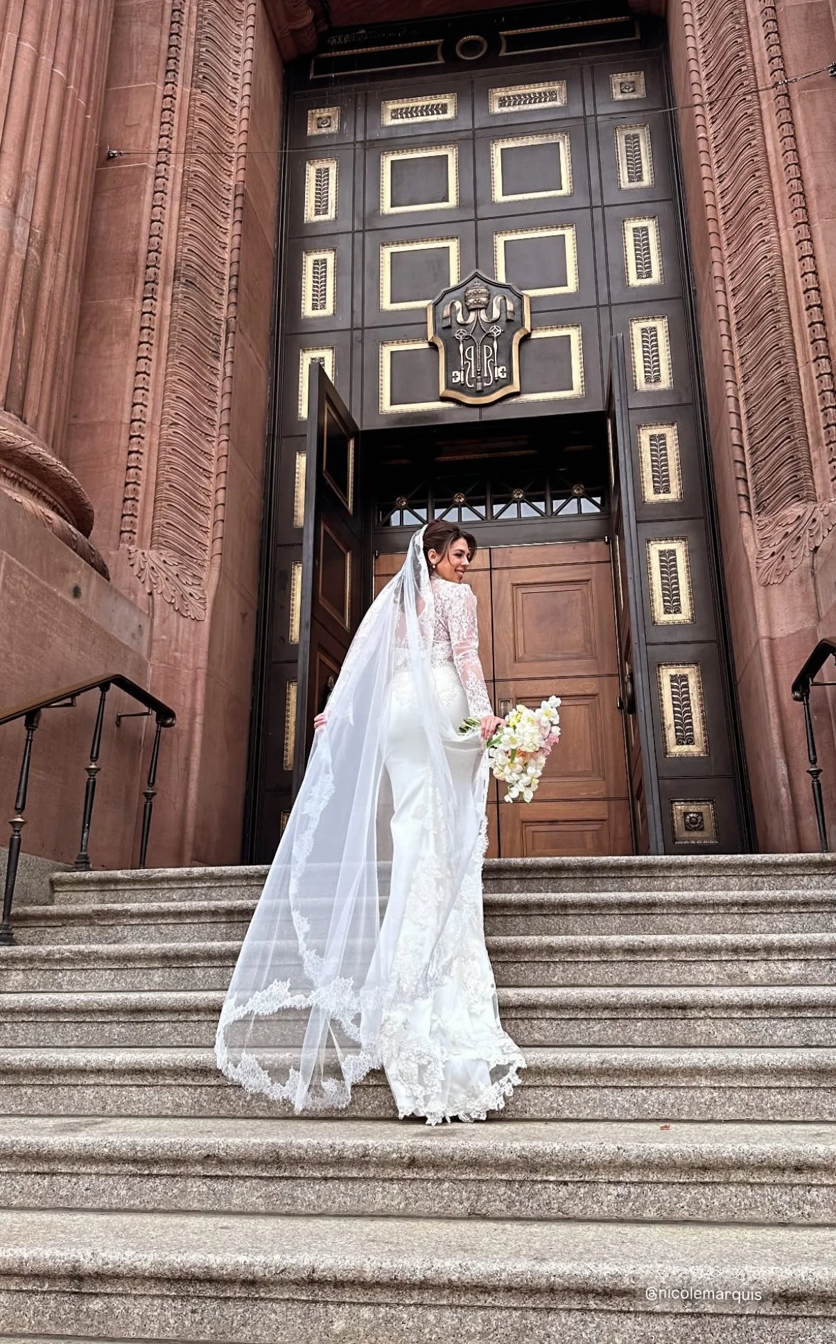 A bride in a white wedding dress with lace details and a long veil, holding a bouquet of white flowers, standing on the steps outside a large historic building with ornate wooden doors and pink stone architecture.