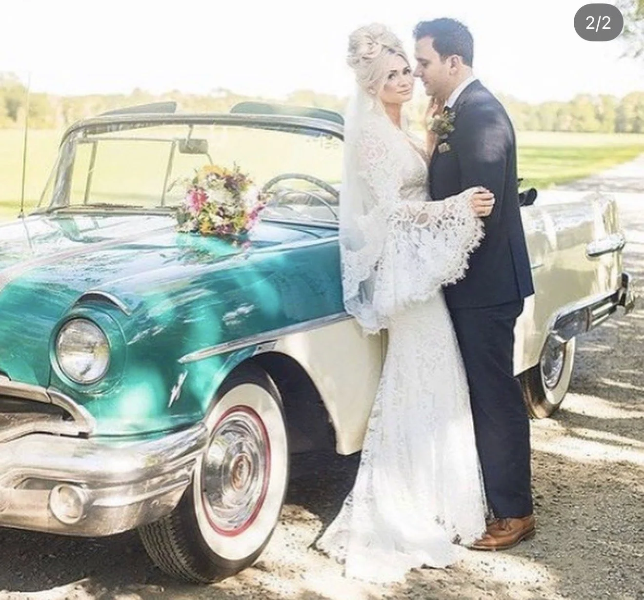A bride and groom stand close together, facing each other, next to a vintage teal and white car decorated with flowers, outdoors on a sunny day.