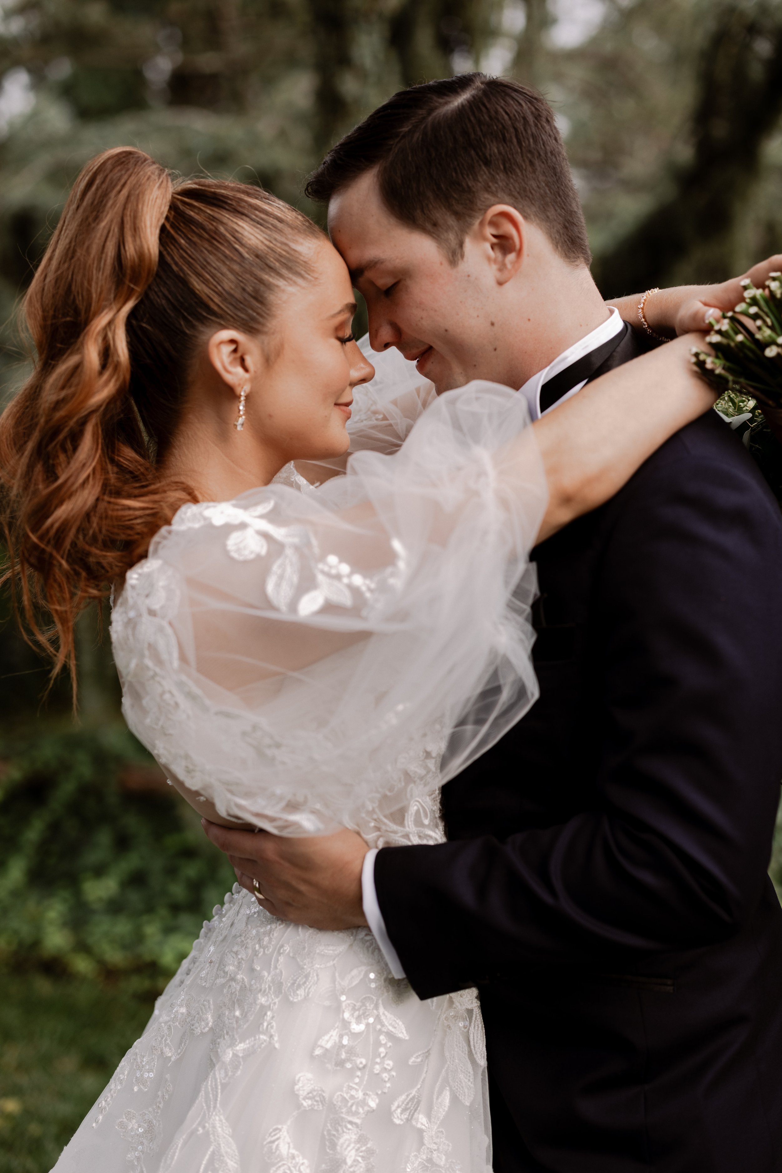 A bride and groom embrace outdoors, their foreheads touching, both with closed eyes, during their wedding. The bride wears a white lace gown with puffed sleeves and the groom a black tuxedo. The background is blurred greenery.