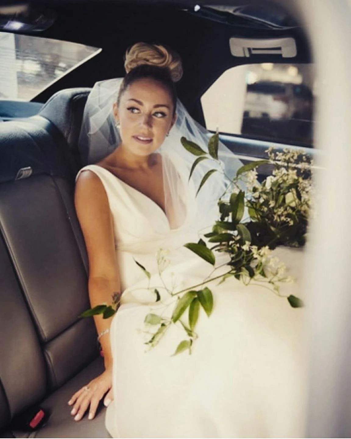 A bride sitting in the backseat of a car, wearing a white wedding dress and veil, with a bouquet of white flowers and green leaves on the table in front of her.