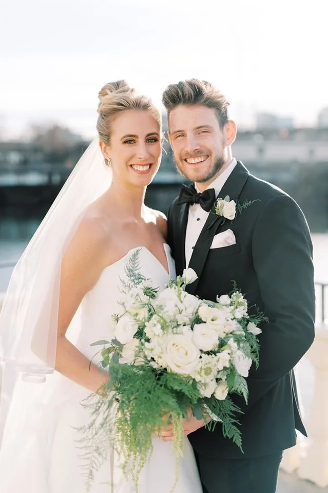 Bride and groom smiling outdoors, bride holding a bouquet of white roses and greenery, bride in a white wedding dress with veil, groom in black tuxedo with bow tie, cityscape in the background.