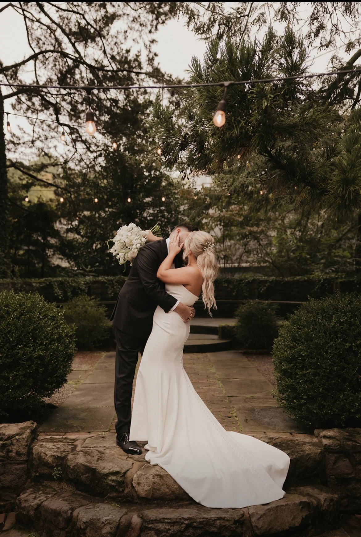 A bride in a white wedding gown and a groom in a black suit kiss on a stone platform in a garden, surrounded by greenery and string lights overhead during sunset.