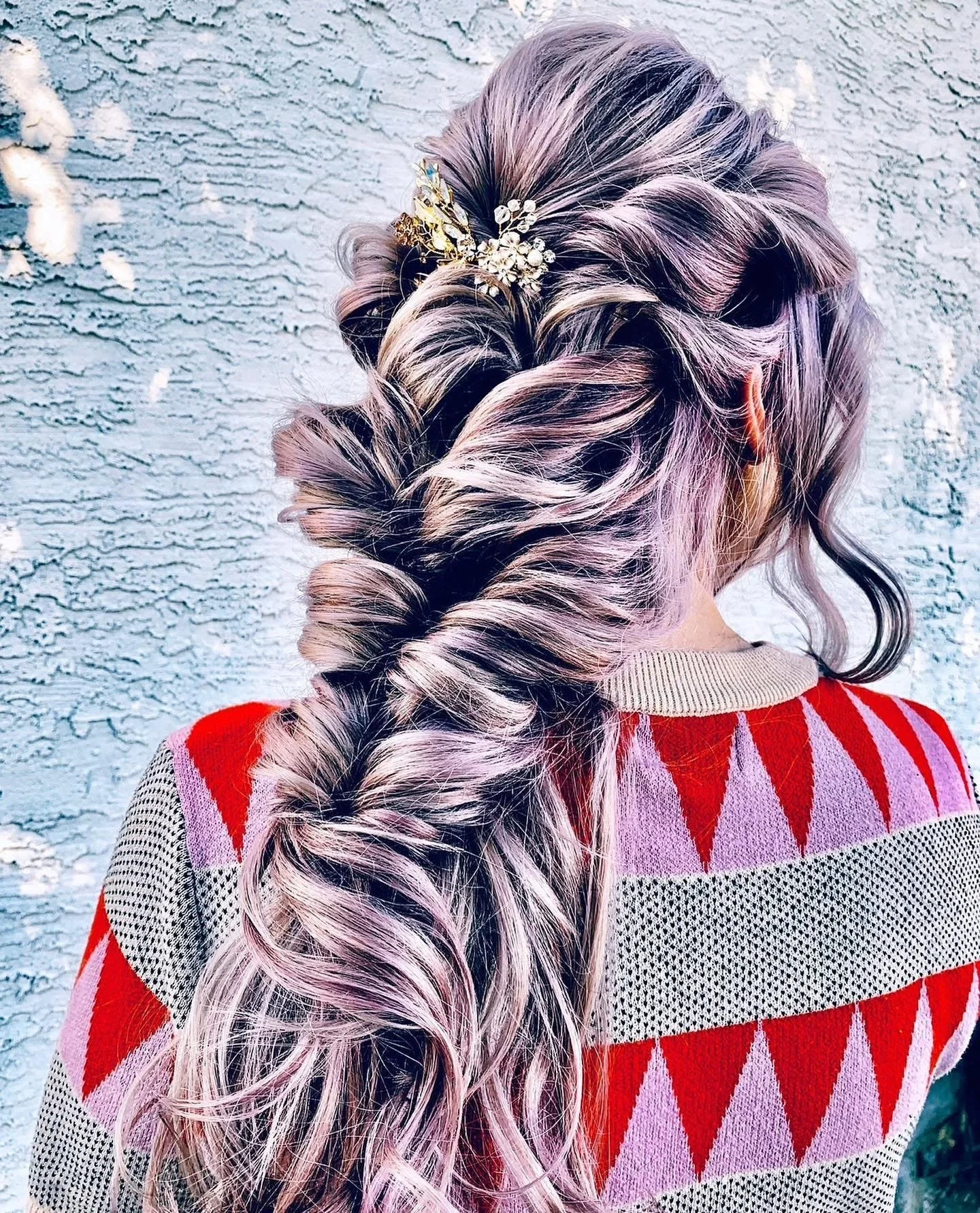 Back of woman with long, wavy hair styled in a braid with a decorative hairpin, wearing a red and beige patterned sweater, standing in front of a textured gray wall.