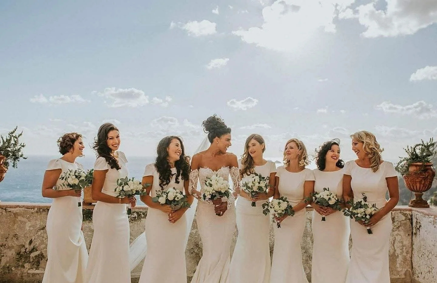 A bride and seven bridesmaids standing outside on a terrace with ocean and sky in the background, all holding bouquets and dressed in white. The bride is in the center, wearing a lace off-the-shoulder wedding gown, and the bridesmaids wear matching s