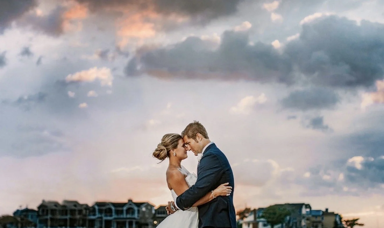 A bride and groom embrace outdoors at sunset, with clouds and residential buildings in the background.