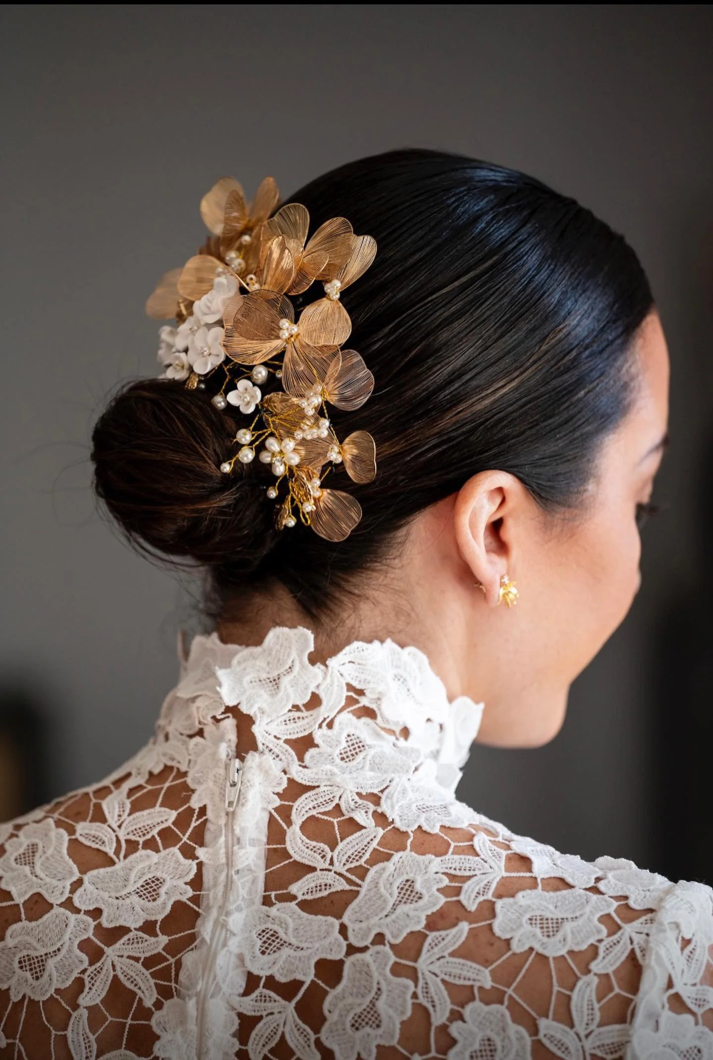 Close-up of a woman's elegant black hair styled in a bun, adorned with a decorative hairpiece featuring gold and white flowers and pearls, wearing a white lace dress with a high collar.