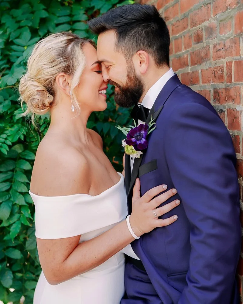 A bride and groom in wedding attire lean their foreheads together, smiling with their eyes closed against a brick wall and green foliage background.