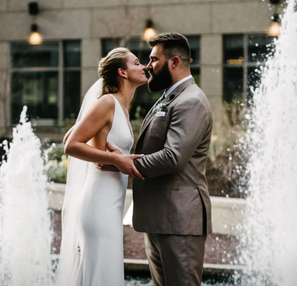 A bride and groom stand close to each other, about to kiss, in front of a fountain, with a building in the background.