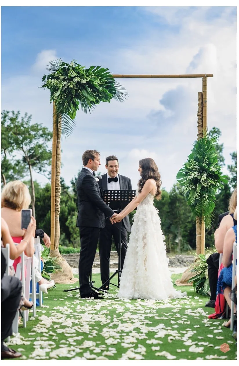 A couple's wedding ceremony outdoors under a bamboo arch decorated with green foliage, with an officiant, guests seated on either side, and petals scattered on the grassy aisle.