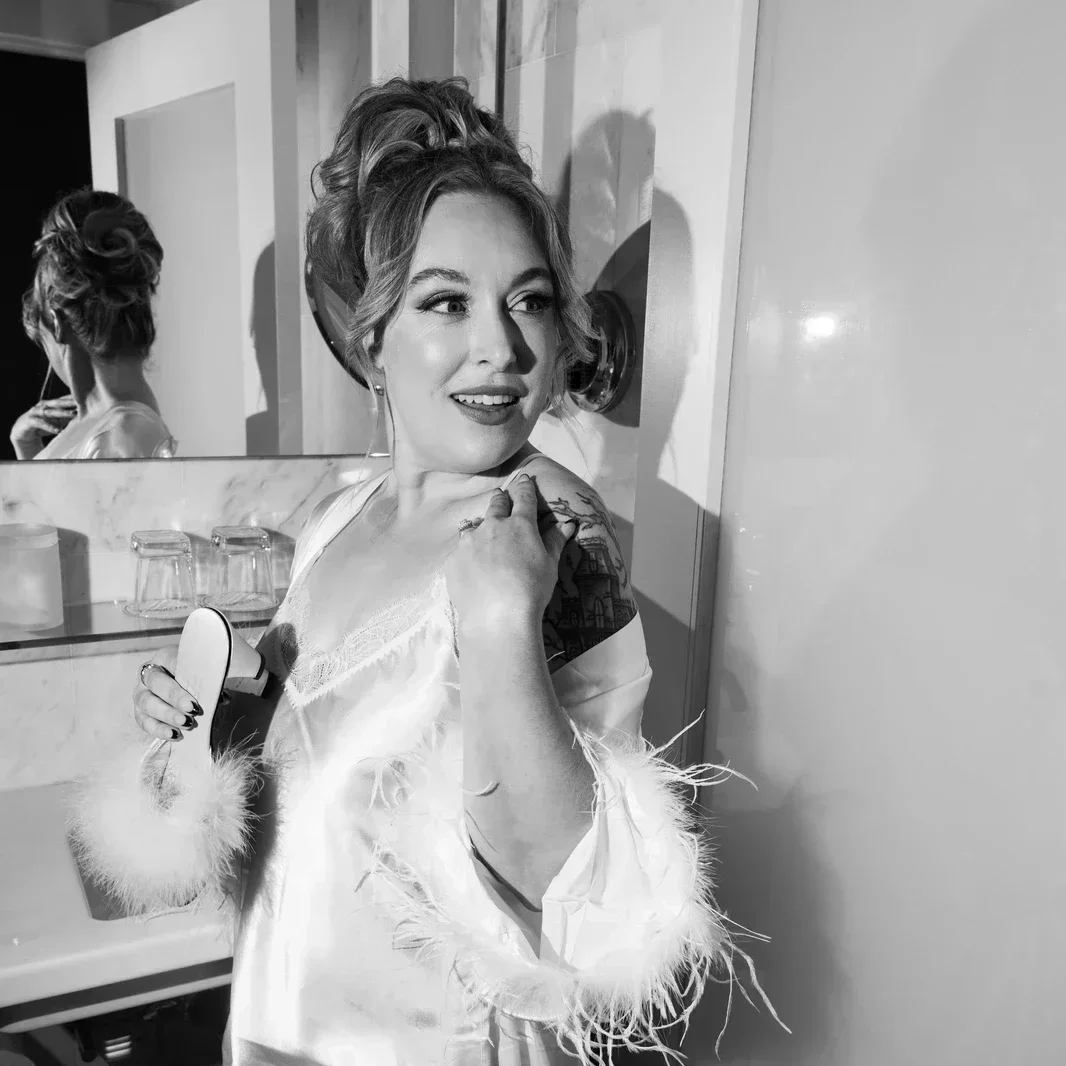 A woman with styled hair, wearing a satin robe with feathered cuffs, smiling and holding a mirror in a bathroom with a mirror reflecting her back, and a wall-mounted hairdryer.