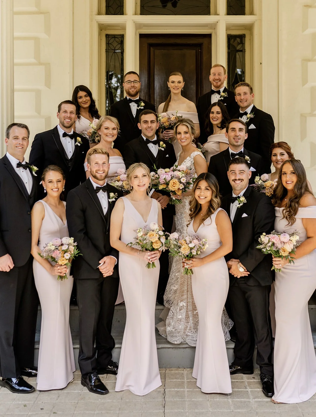 Group of wedding party members, including bridesmaids in white dresses and groomsmen in black tuxedos, standing on stairs in front of a building with decorative windows, holding bouquets of pastel flowers.