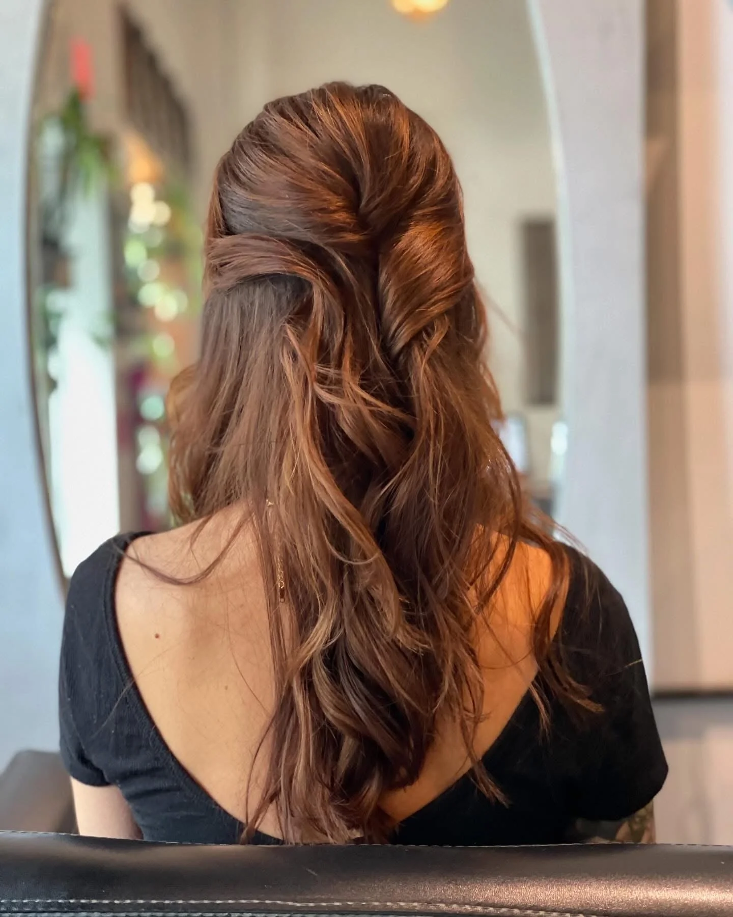 A woman with long, reddish-brown hair styled in loose curls, turned away from the camera, wearing a black top with an off-the-shoulder design, in front of a mirror in a room with soft lighting and decorative plants in the background.