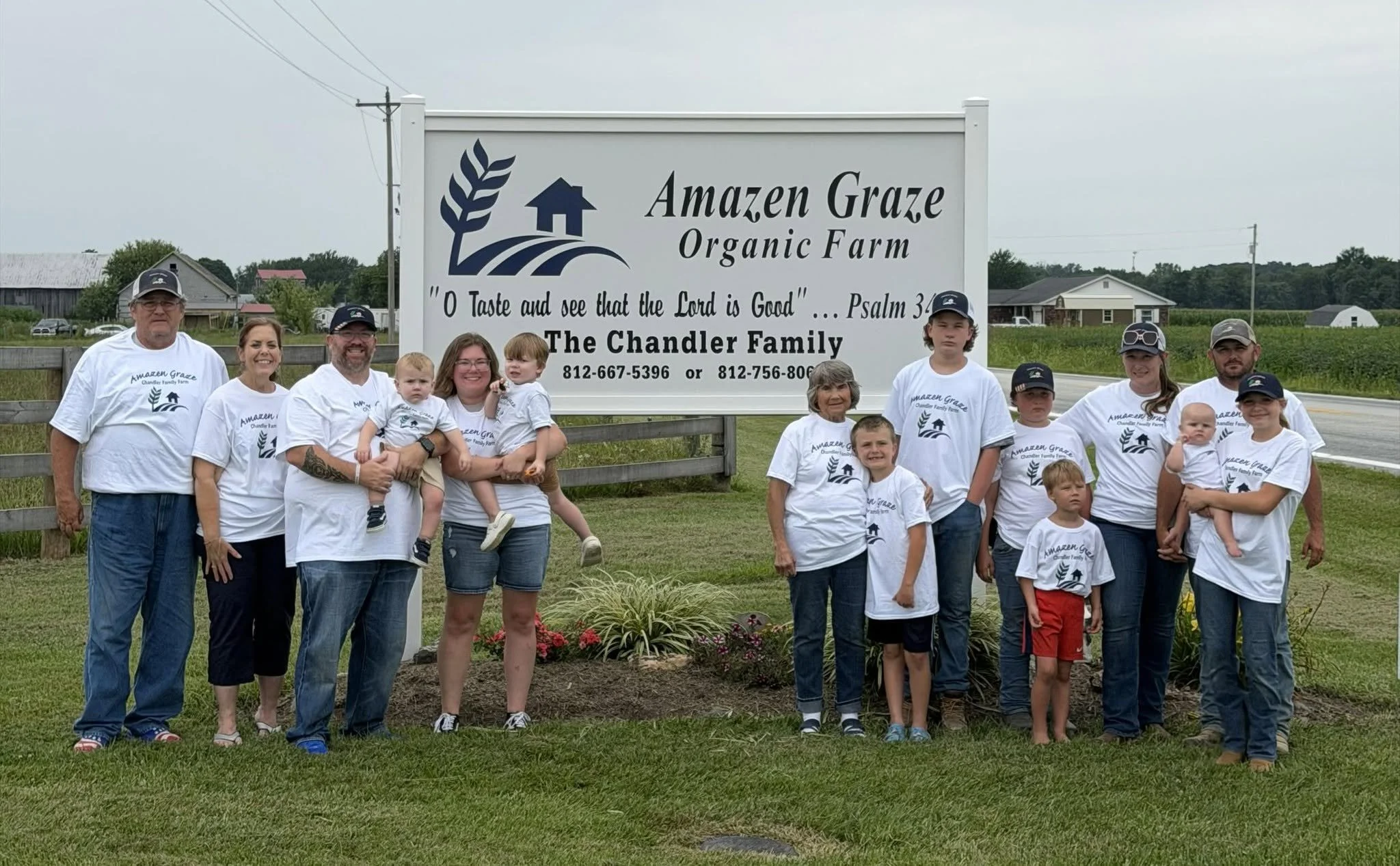 A group of people, including children, standing in front of a large sign that reads 'Amazen Graze Organic Farm' with a quote from Psalm 34. They are wearing white shirts with the farm's logo, and the background shows houses, a field, and a cloudy sky.