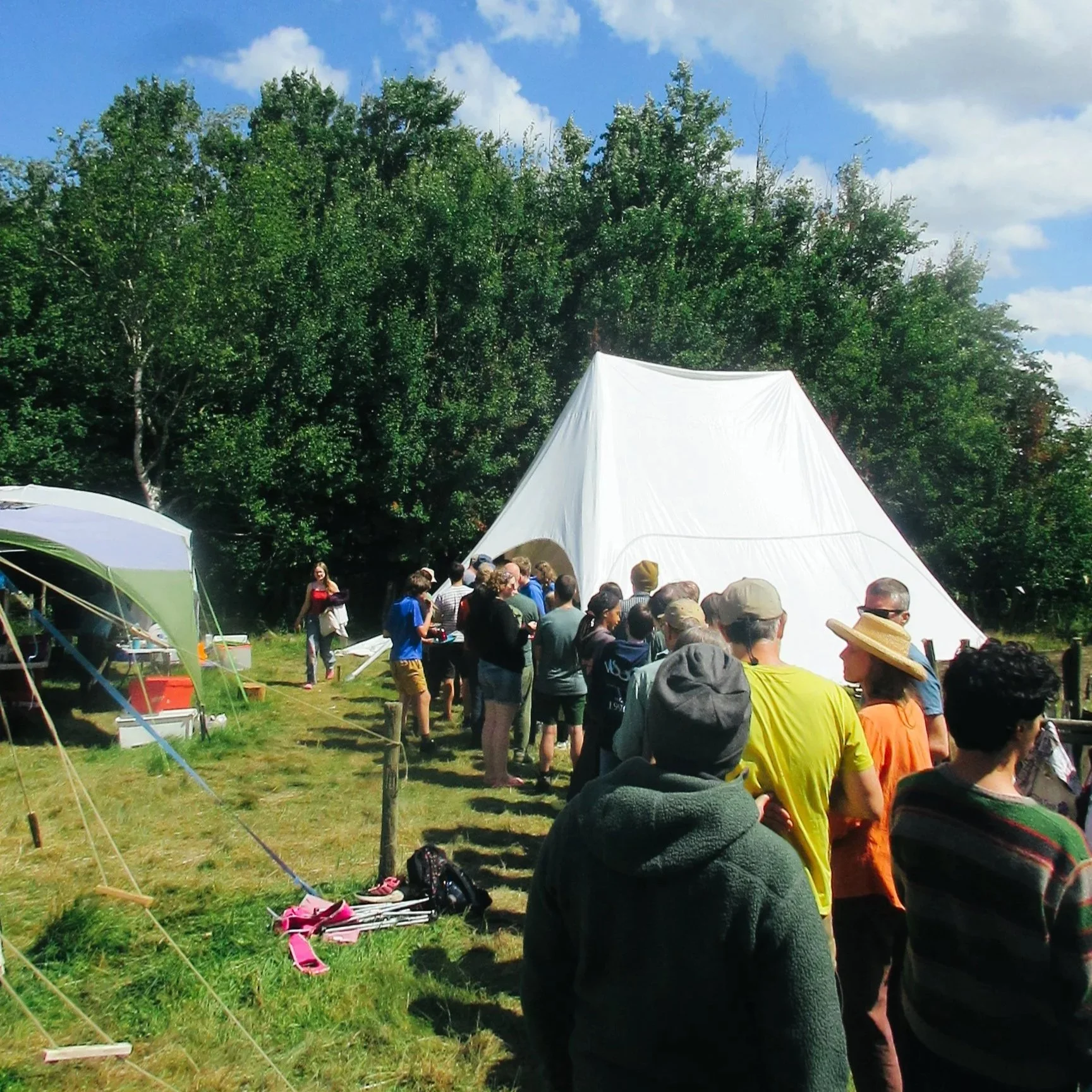Crowd of people standing in line outdoors near a large white tent with trees and blue sky in the background.
