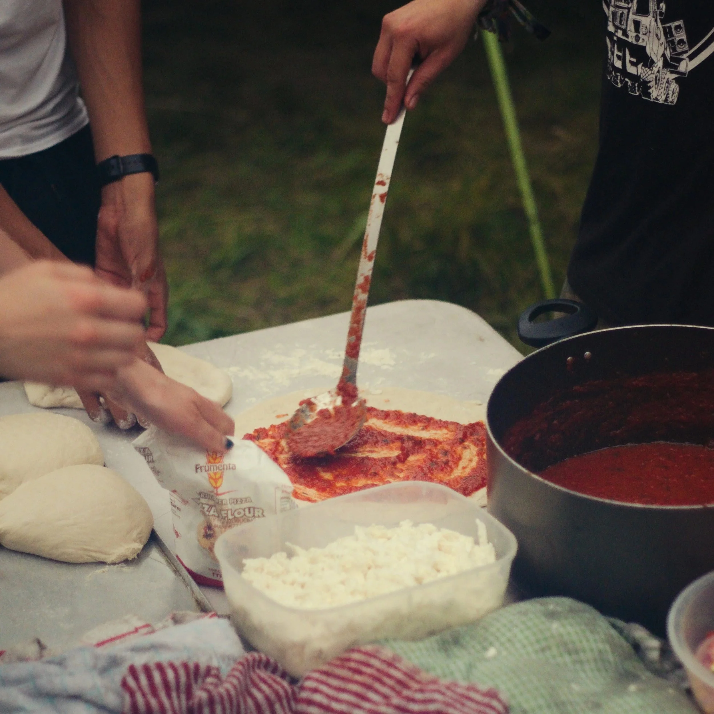 Close-up of a person slicing pizza with a pizza cutter, with various ingredients and containers of cheese and tomato sauce for making pizza in the background.