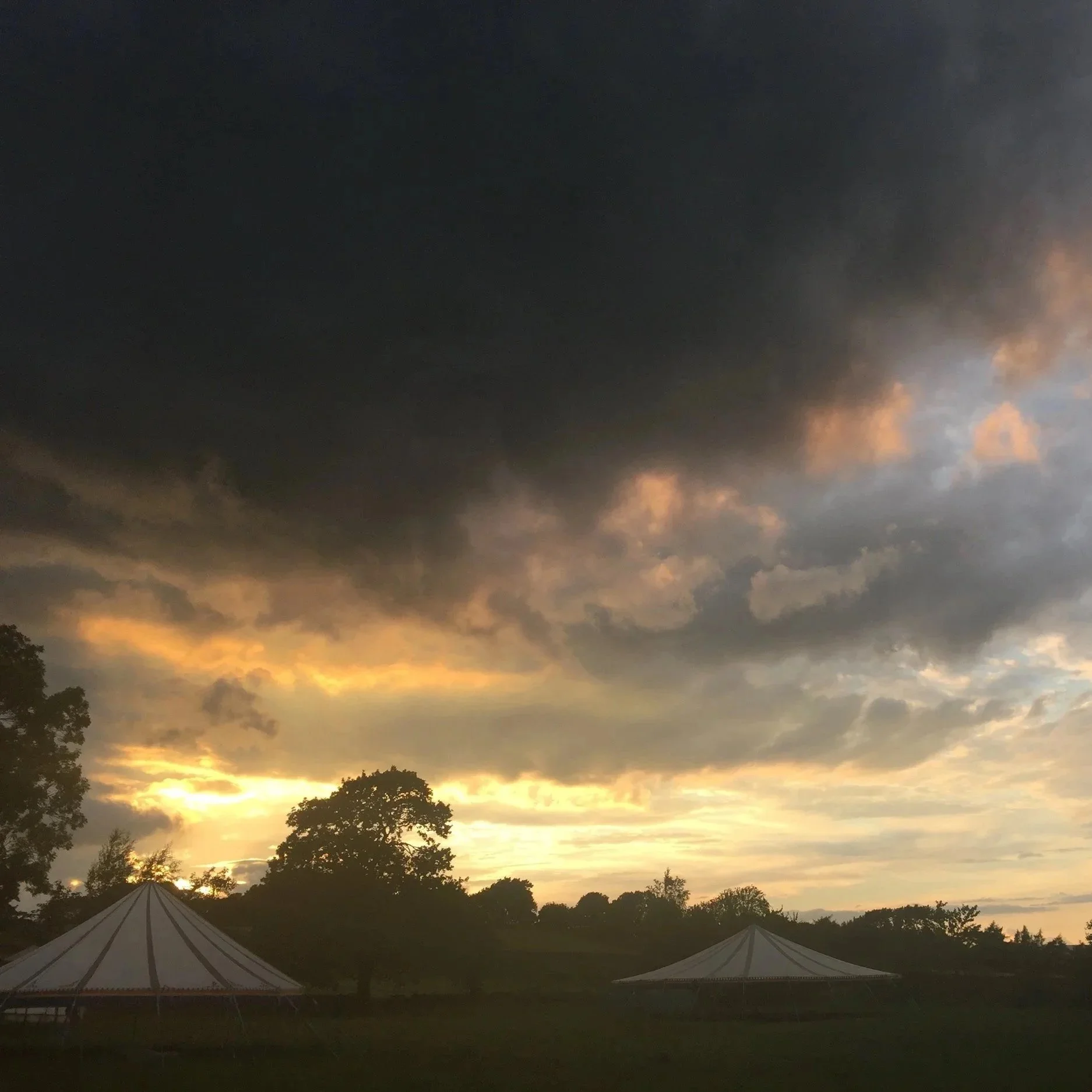 A scenic view of a sunset with a sky filled with dark clouds and patches of orange and yellow light near the horizon. Silhouettes of trees and tents are visible in the foreground.