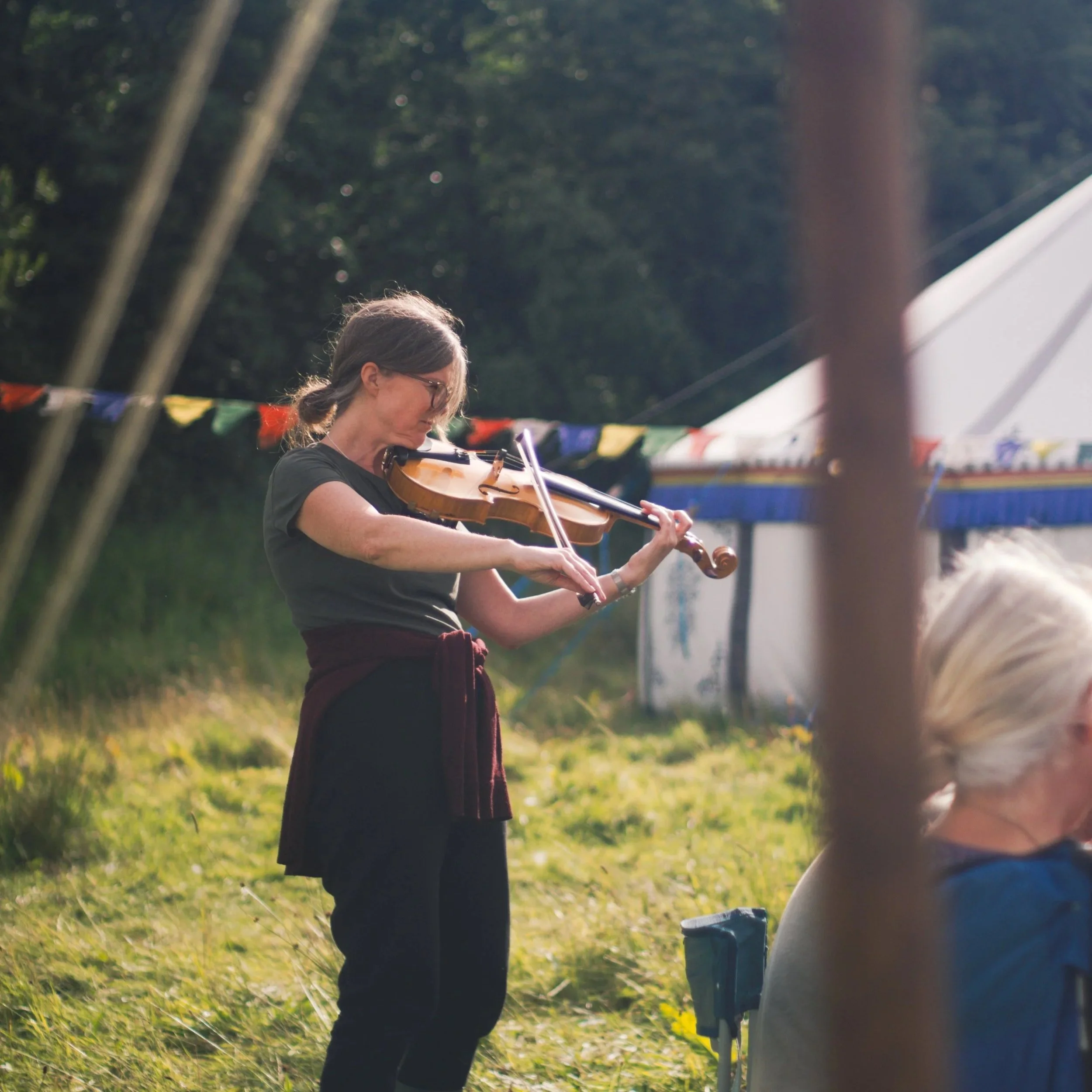 A woman with gray hair and glasses playing the violin outdoors at a park or fairground, with colorful flags hanging in the background, during daytime.