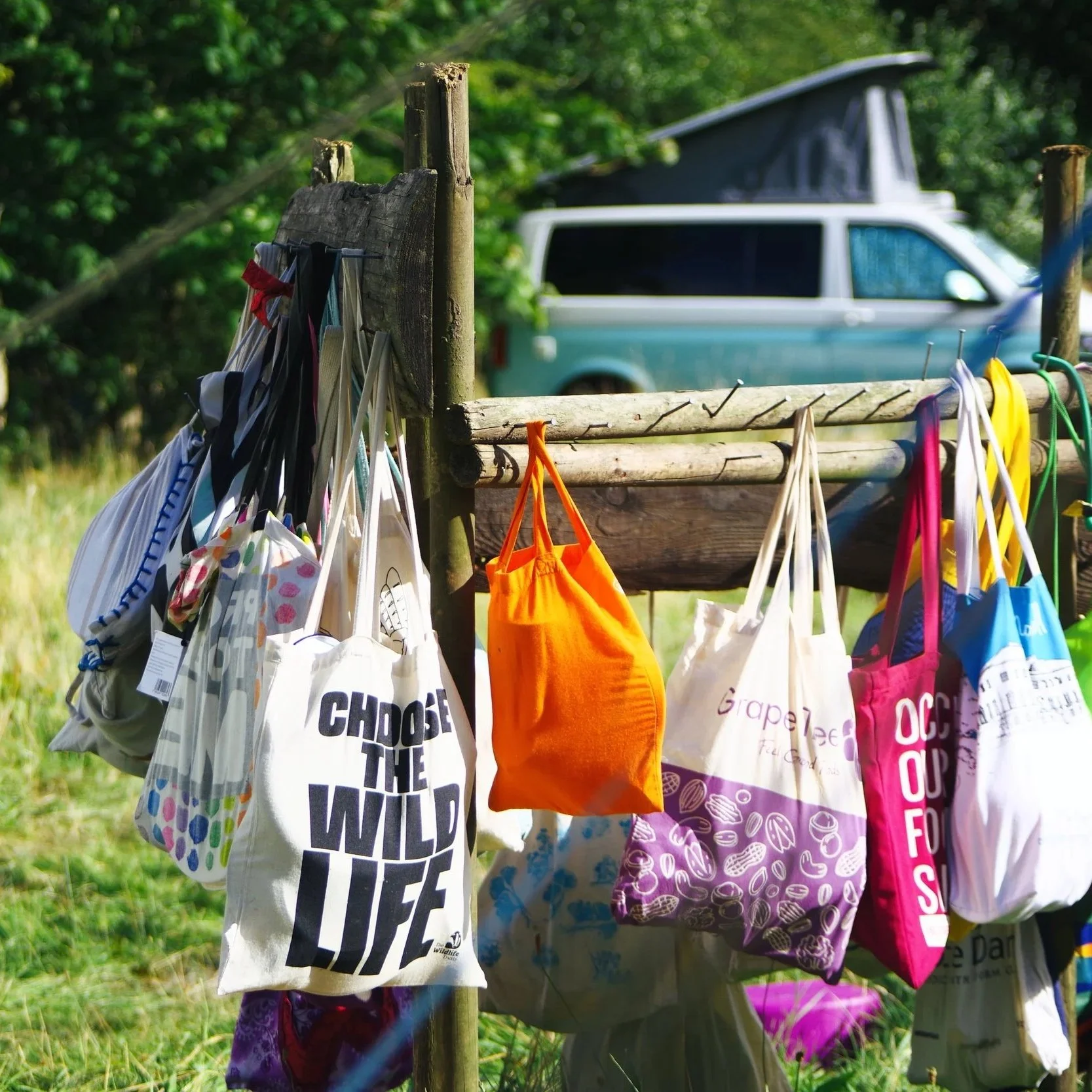 Colorful reusable shopping bags hanging from a wooden fence post outdoors, with a camper van in the background surrounded by greenery.
