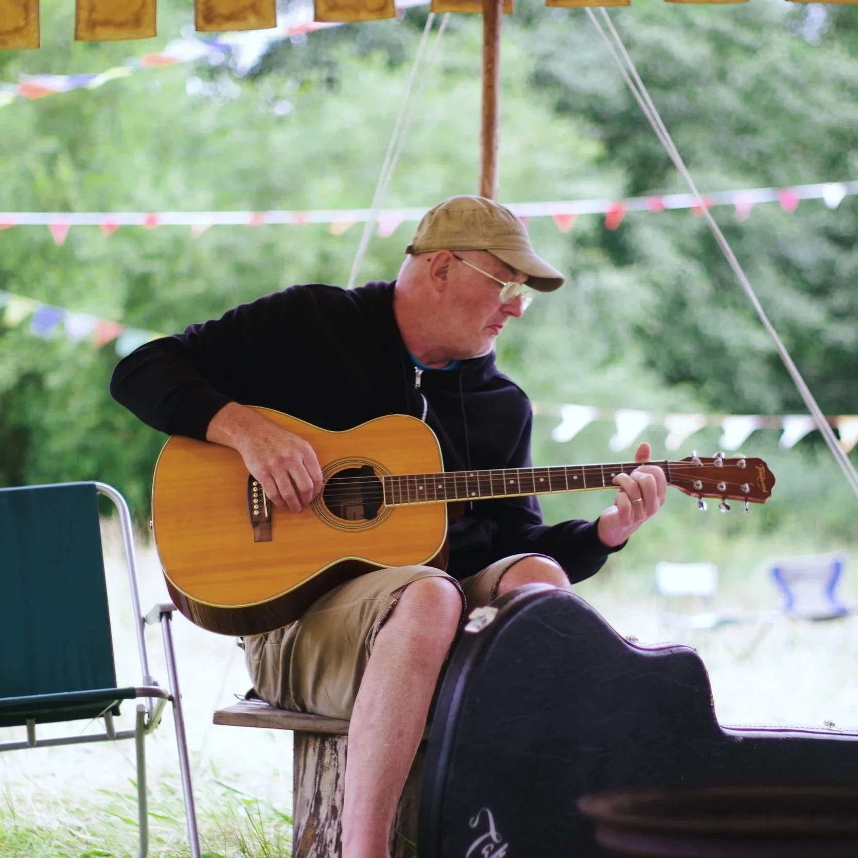 A man wearing a tan cap glasses and black hoodie sitting outdoors playing an acoustic guitar on a bench with a natural background and other chairs nearby.