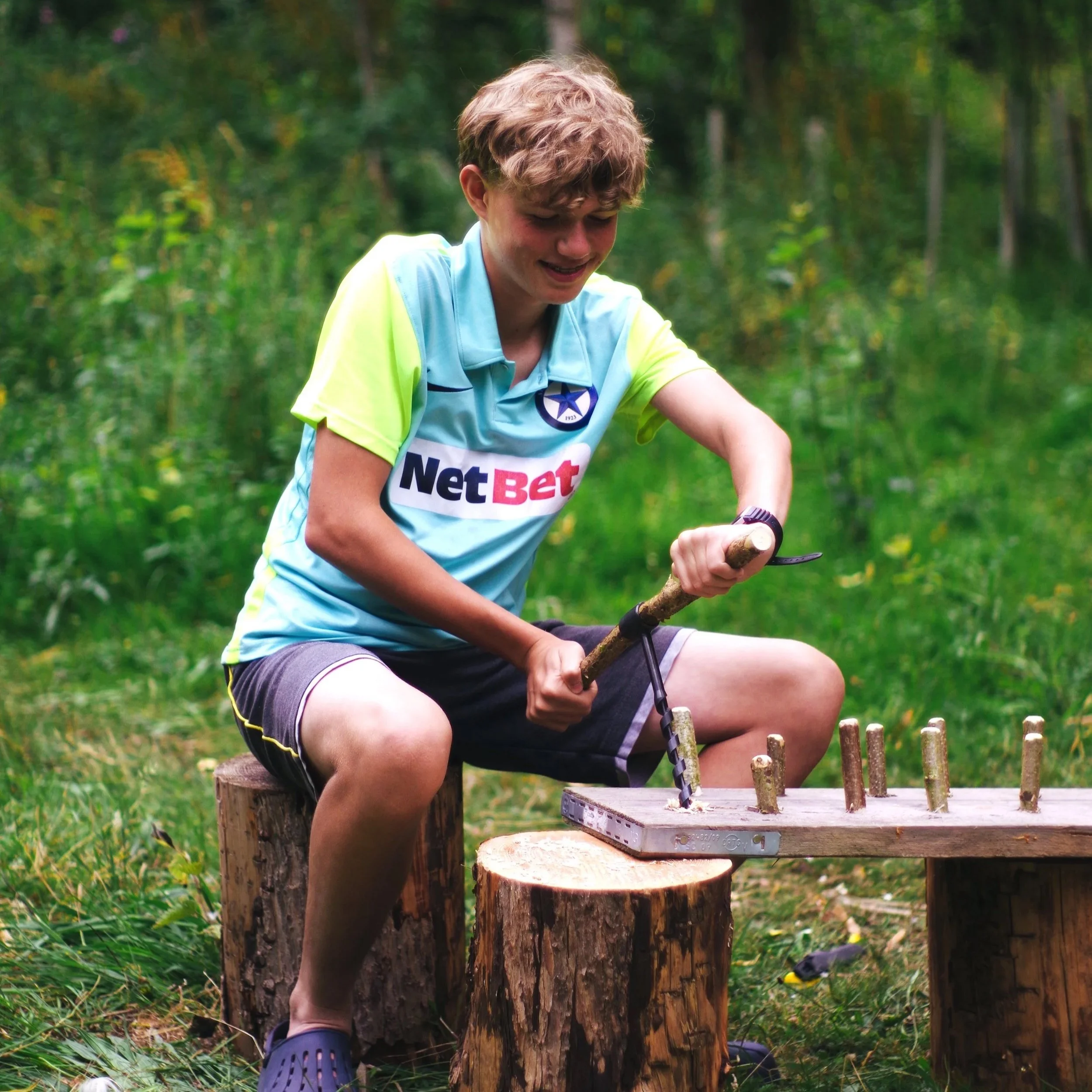 Boy sitting on a tree stump in a forest, holding a tool and smiling, wearing a light blue sports shirt with a 'NetBet' logo, shorts, and Crocs.