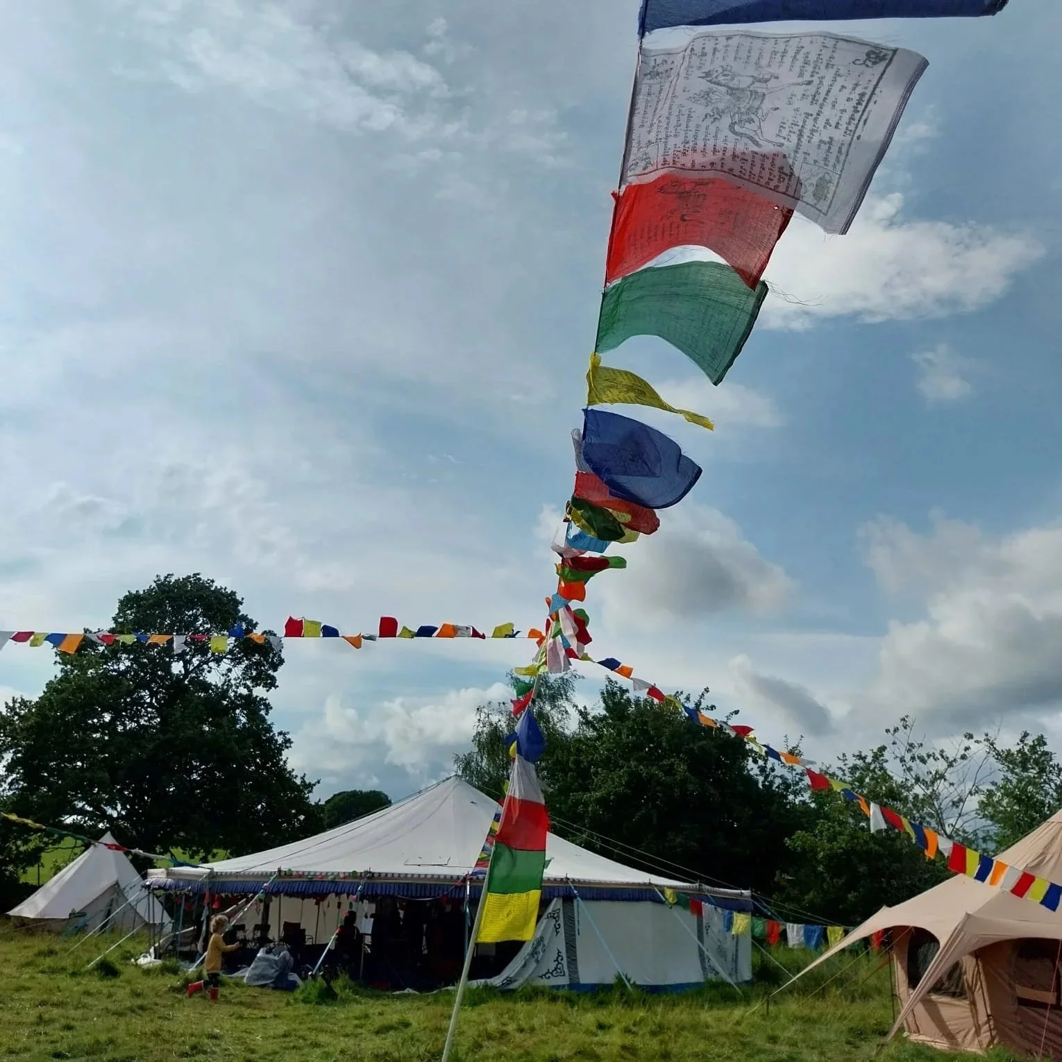 Colorful flags hanging above a festival tent set up outdoors, with trees and cloudy sky in the background.