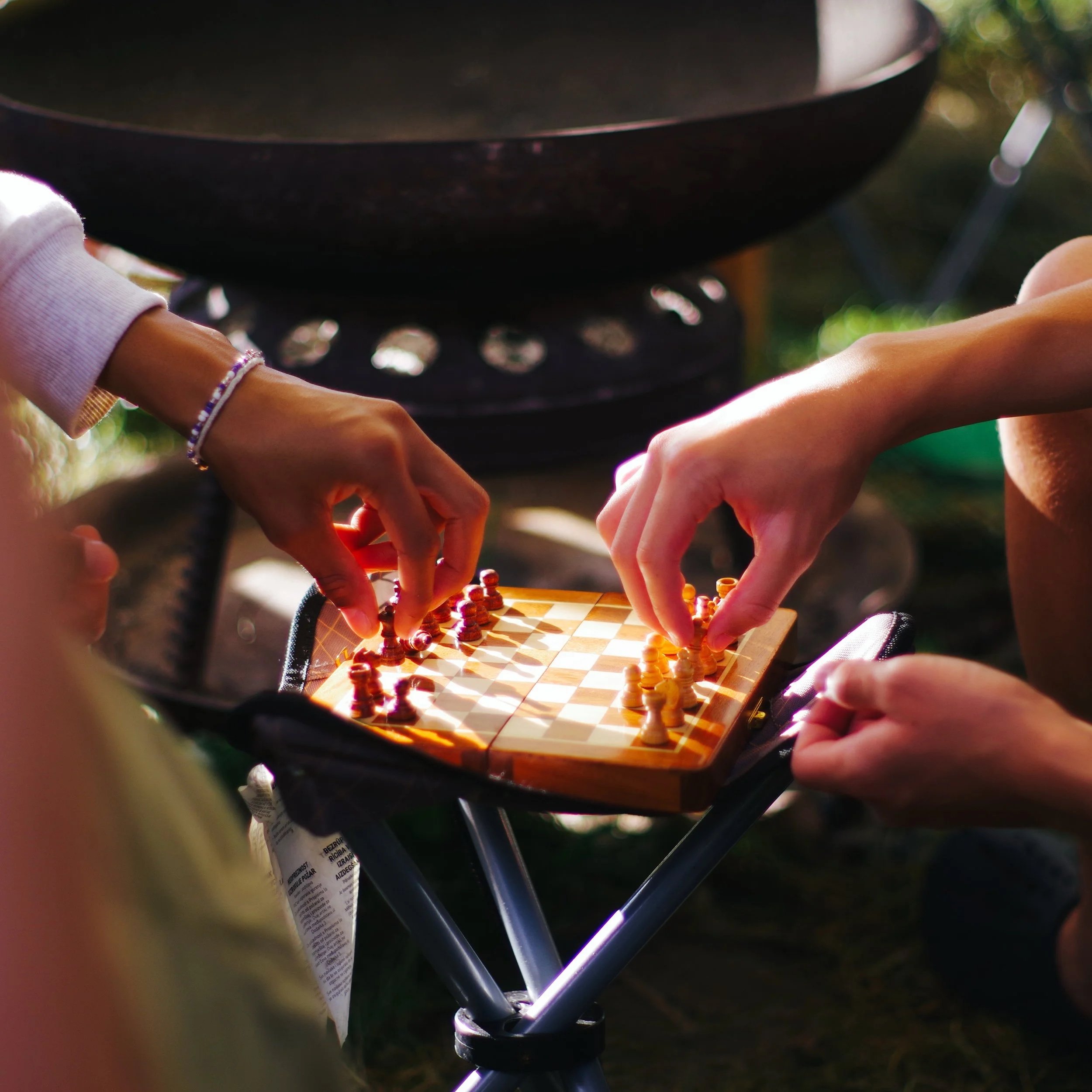 Close-up of a chess game being played outdoors, focused on the chessboard and hands of players, with a black barbecue grill in the background and natural outdoor lighting.