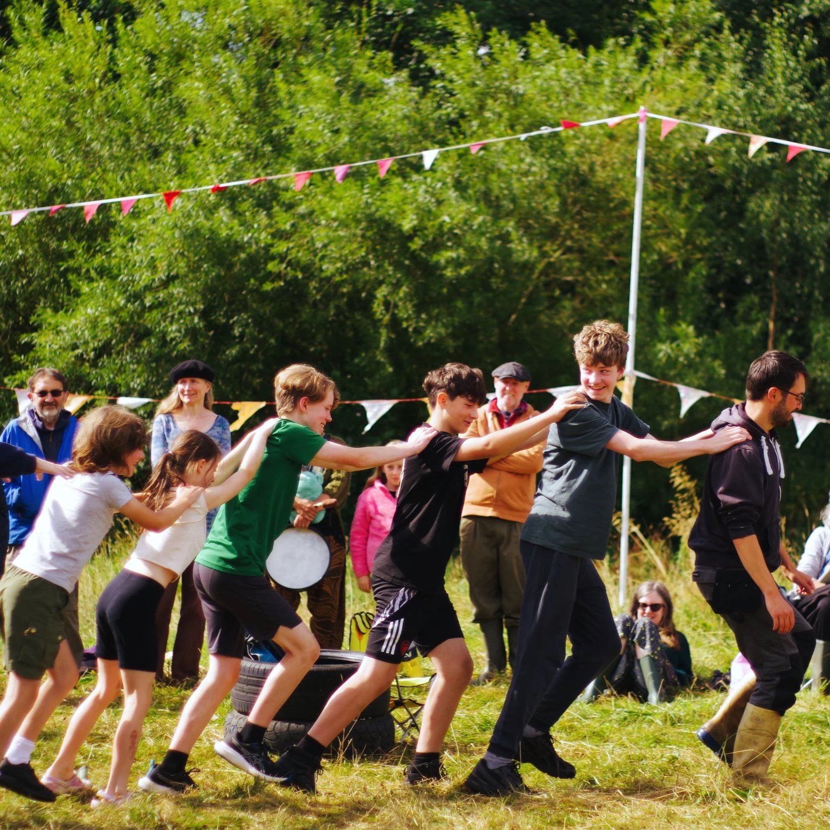 People participating in a tug of war game outdoors, with trees and bunting flags in the background.