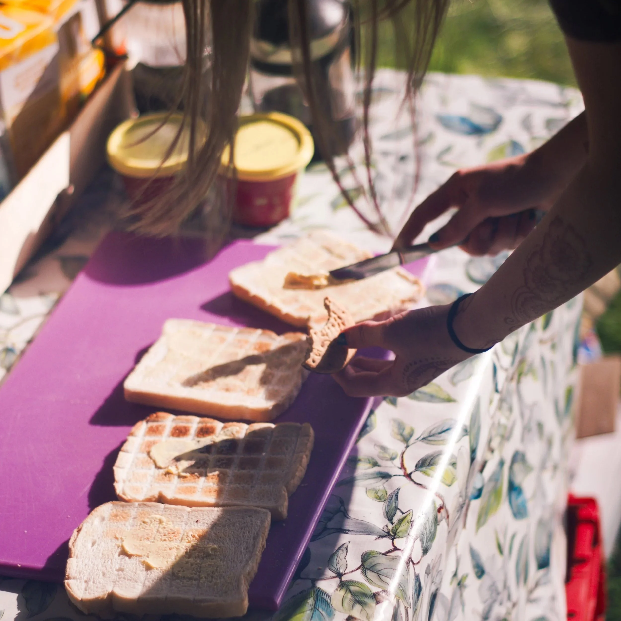 A close-up of slices of bread on a purple cutting board outdoors, with a person’s hand using tongs to handle the bread. There is a yellow and brown container in the background, and the scene is set on a table covered with a floral tablecloth.