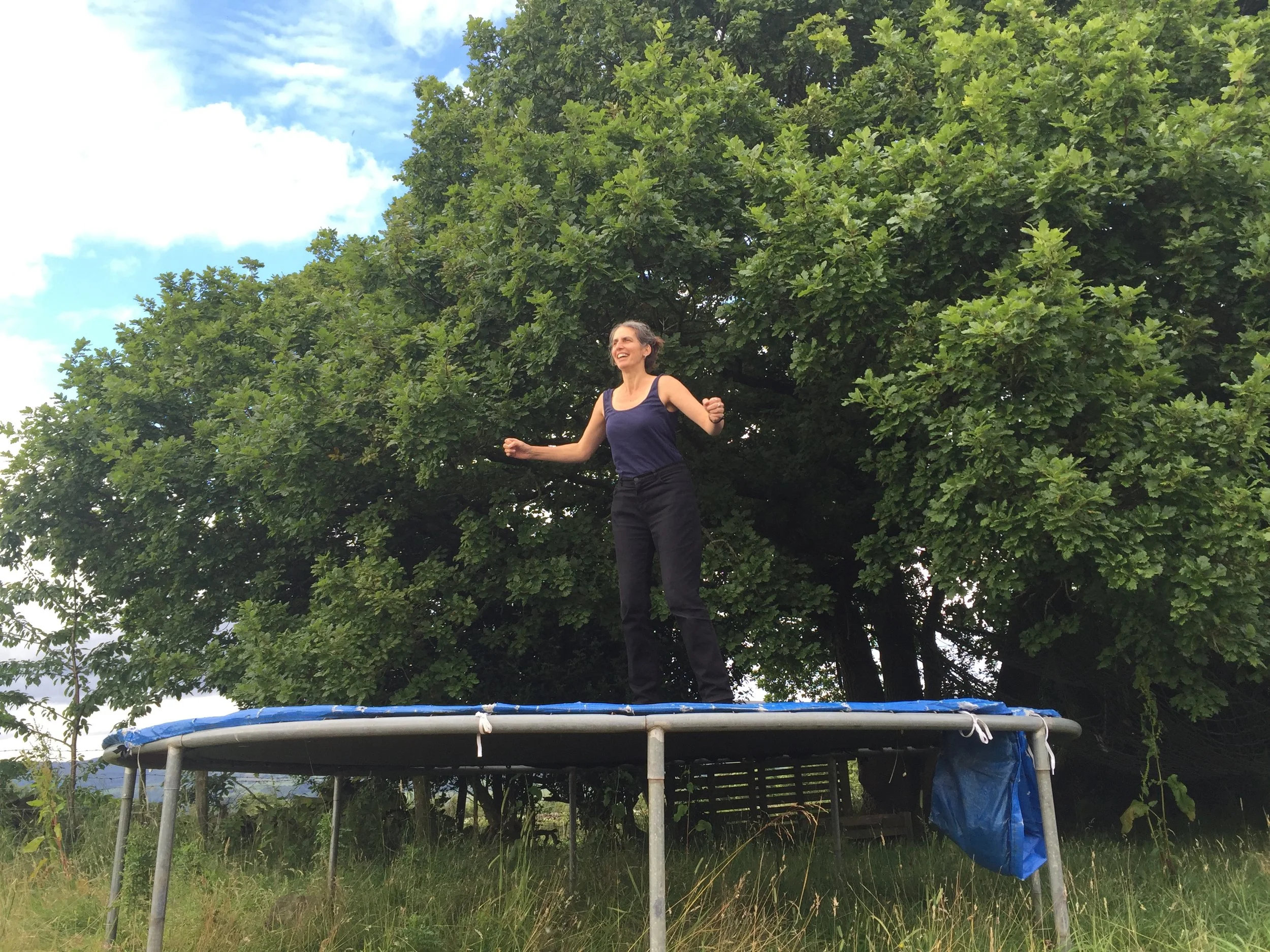 A woman smiling while standing on a trampoline outdoors under a large leafy tree.