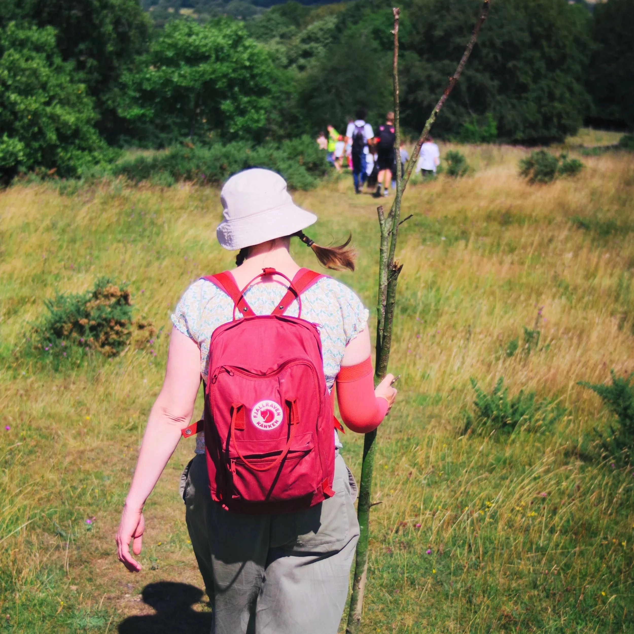 Child with a white hat and red backpack walking on a trail through a green field with trees and hills in the background.