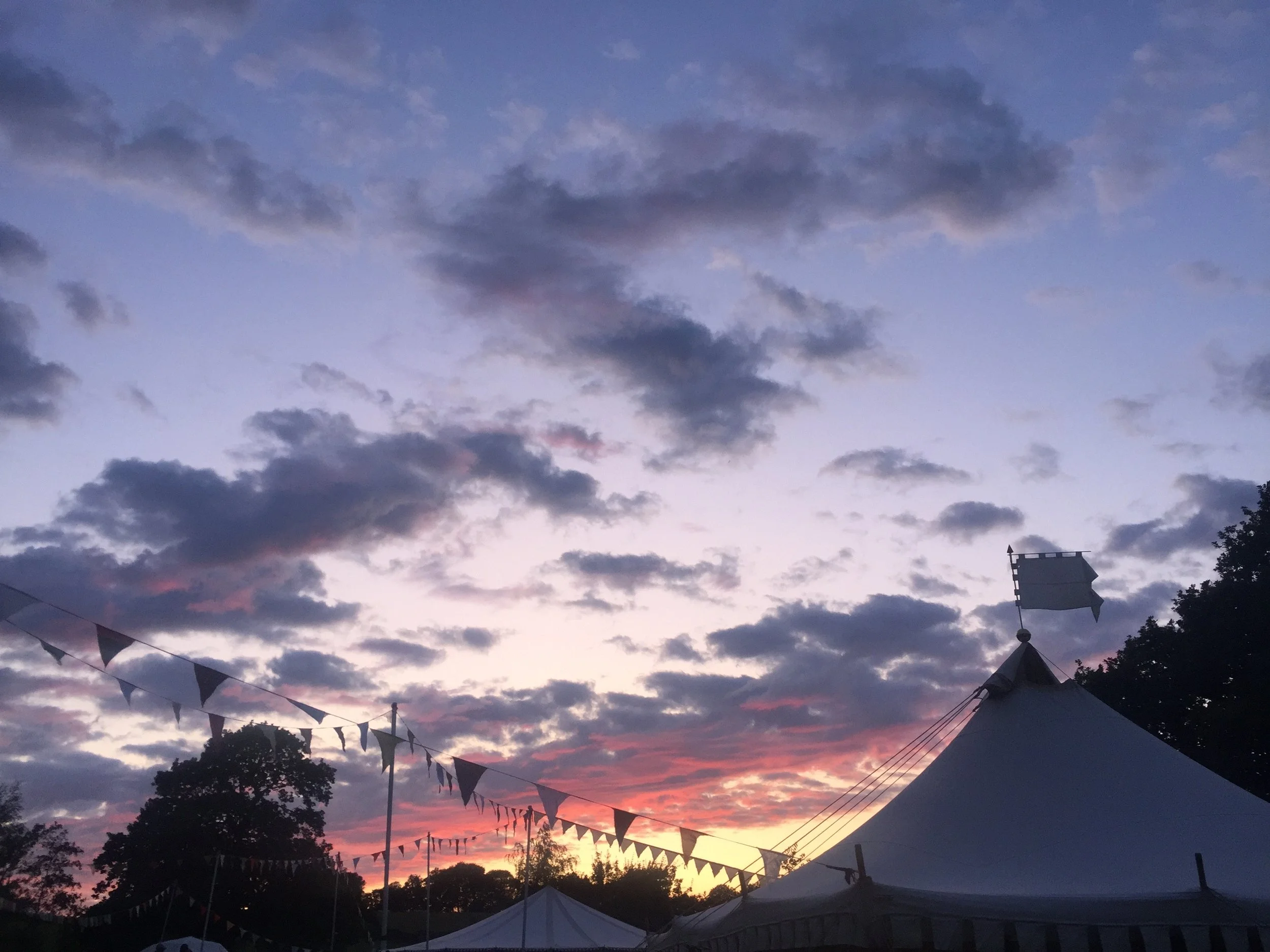 A sky during sunset filled with dark and pink clouds, with decorative flags and tents in the foreground, and trees at the bottom.