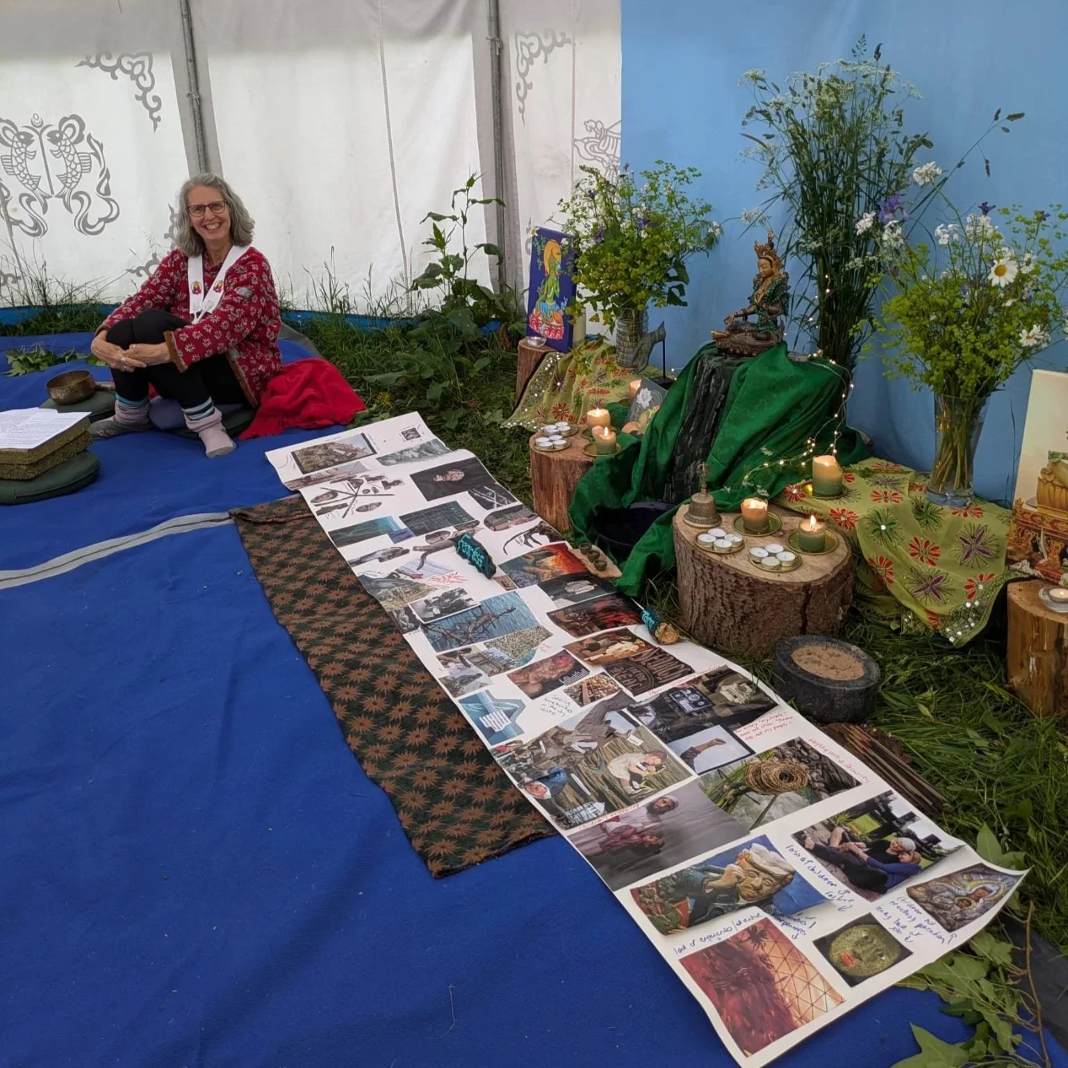 A woman with gray hair and glasses sitting on a blue mat inside a decorated tent, with a display of photographs, artwork, candles, and plants on the ground next to her.