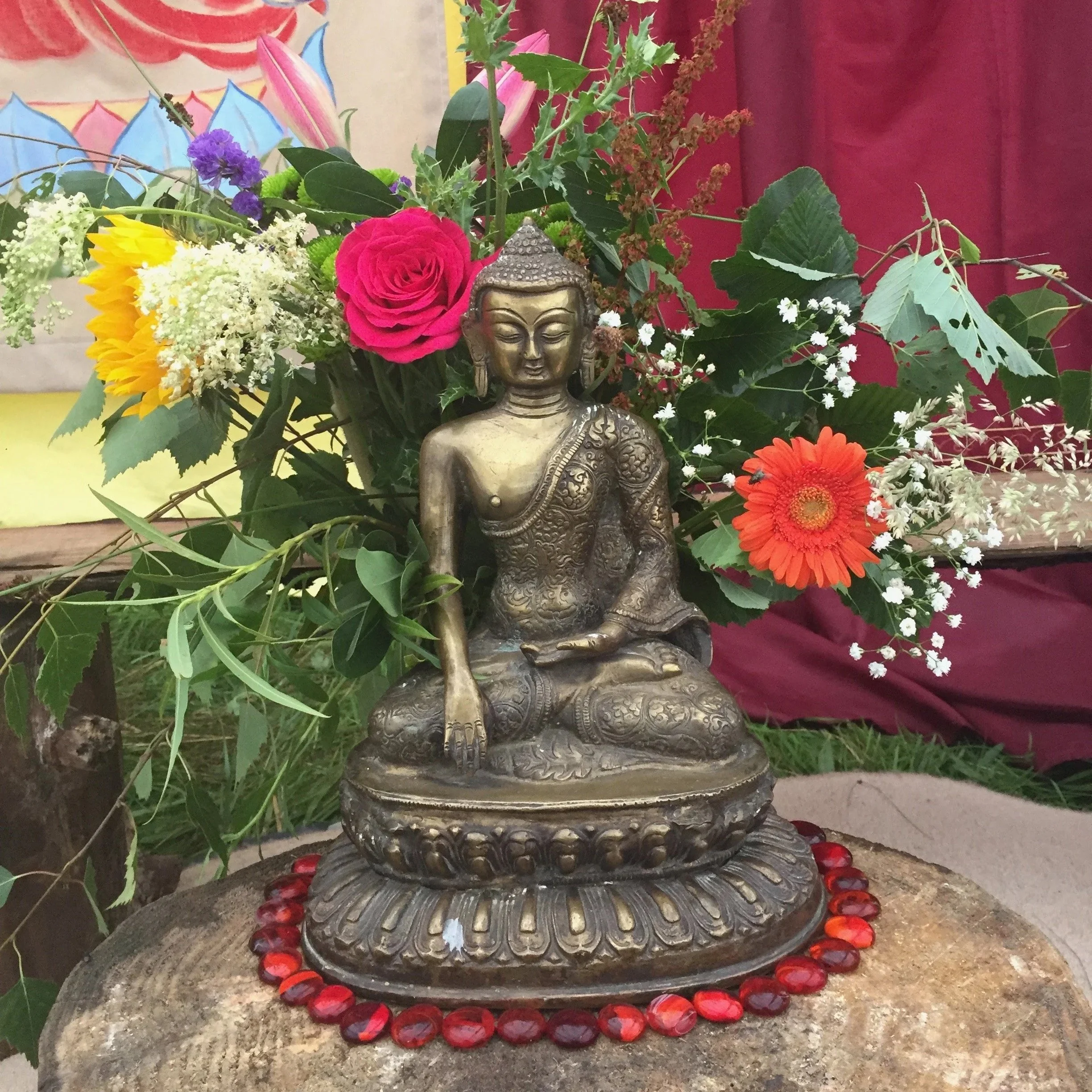 A metal Buddha statue surrounded by colorful flowers including pink roses, orange gerbera daisies, and various greenery, with a red curtain in the background.