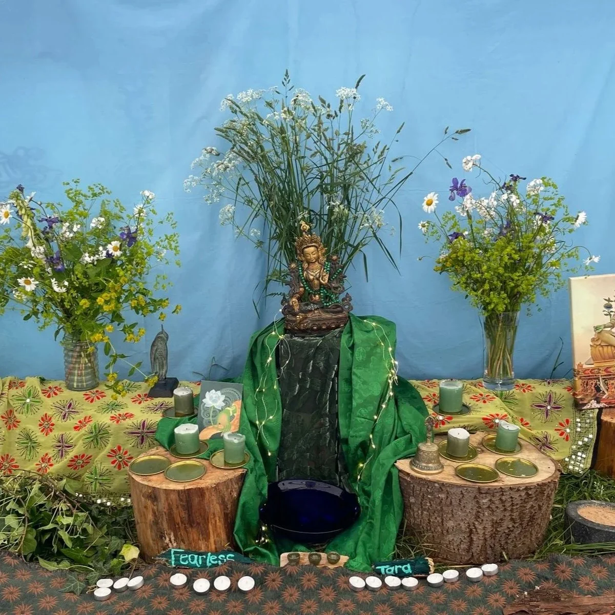 An altar decorated with green drapes, candles, and greenery, with a statue of a deity at the center, surrounded by flowers and wood stumps used as tables, on a floral tablecloth against a blue background.