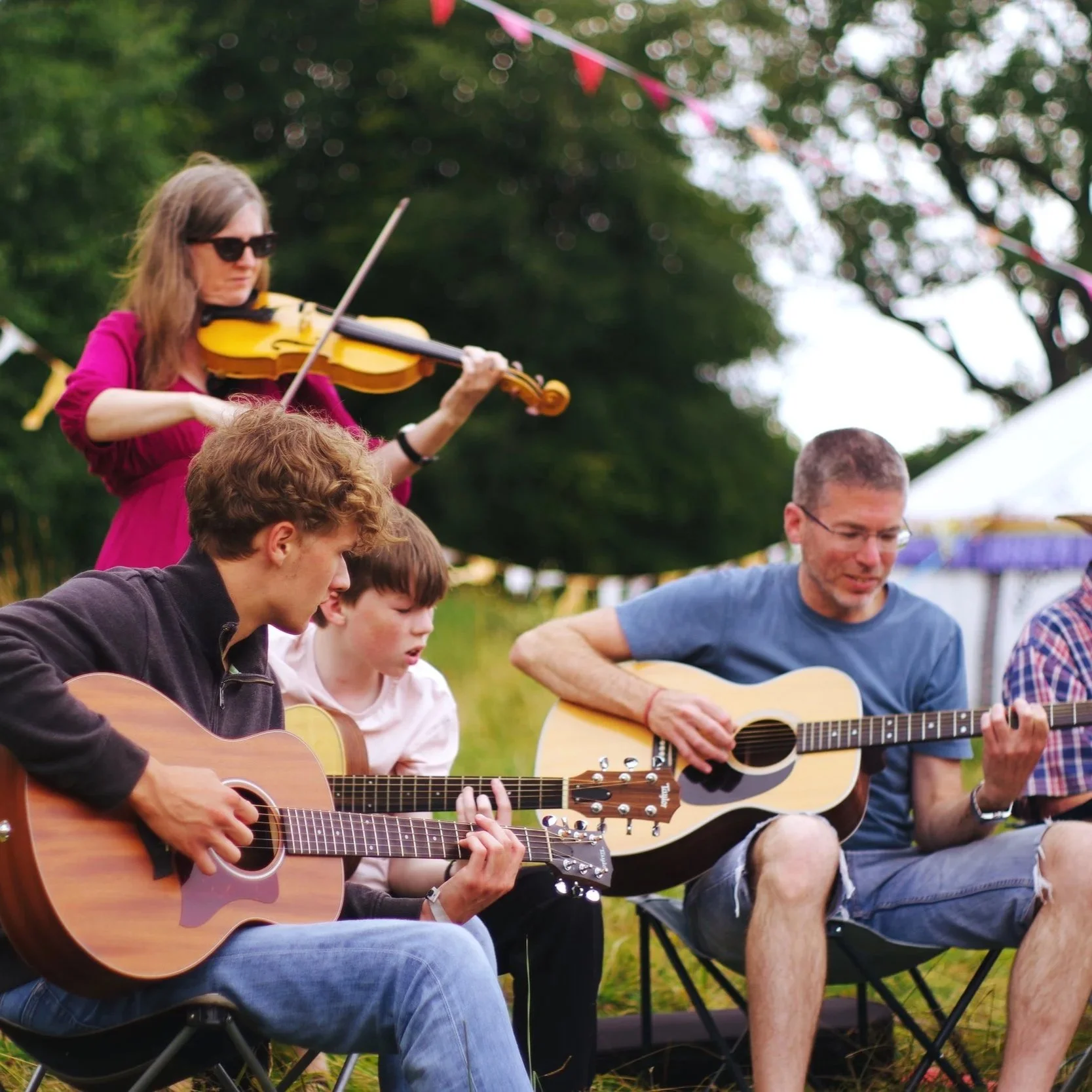 A group of people sitting outdoors playing guitars and singing at a festival or gathering with trees and bunting in the background.