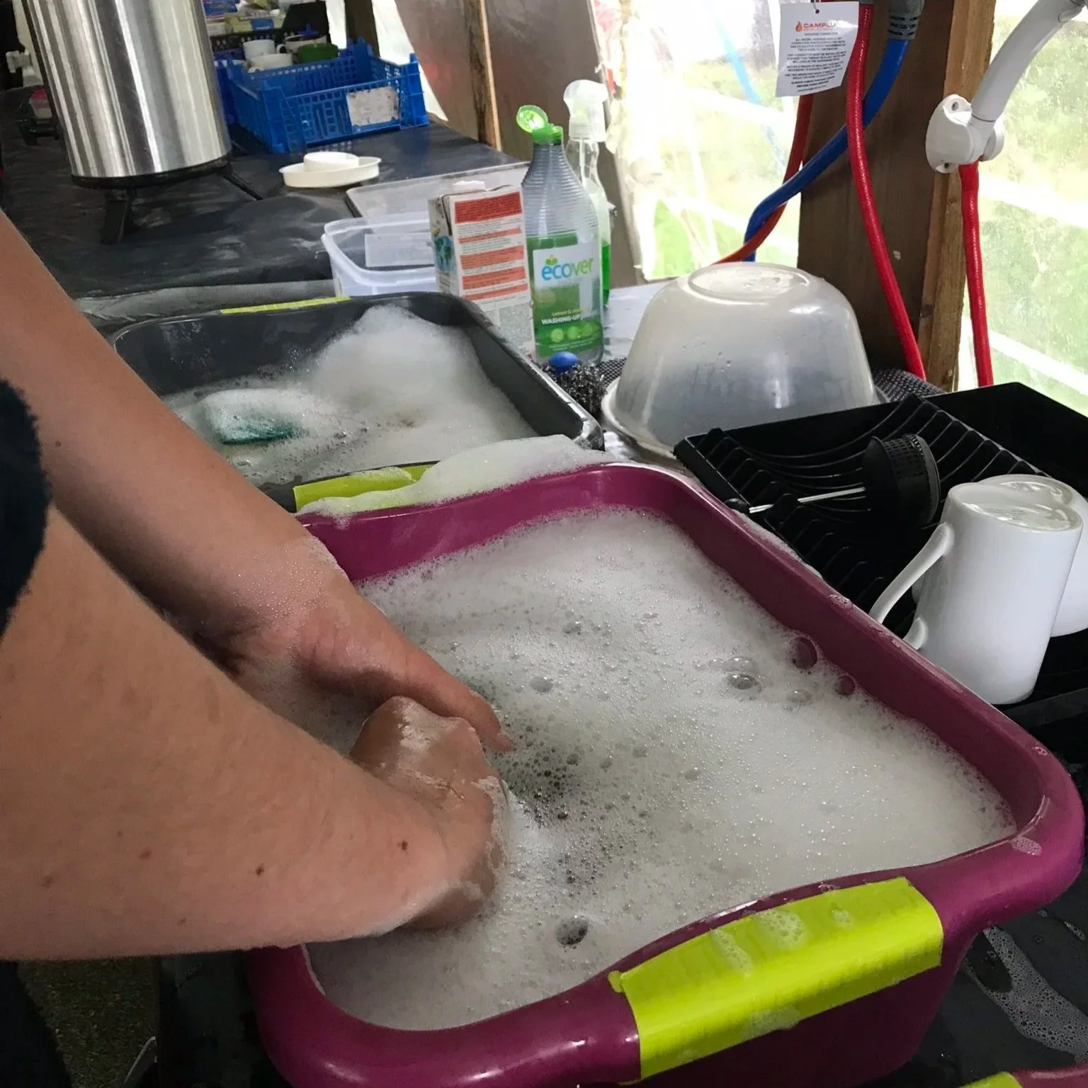 Person washing dishes in a pink plastic tub filled with soapy water, with various kitchen items and cleaning supplies on the counter nearby.