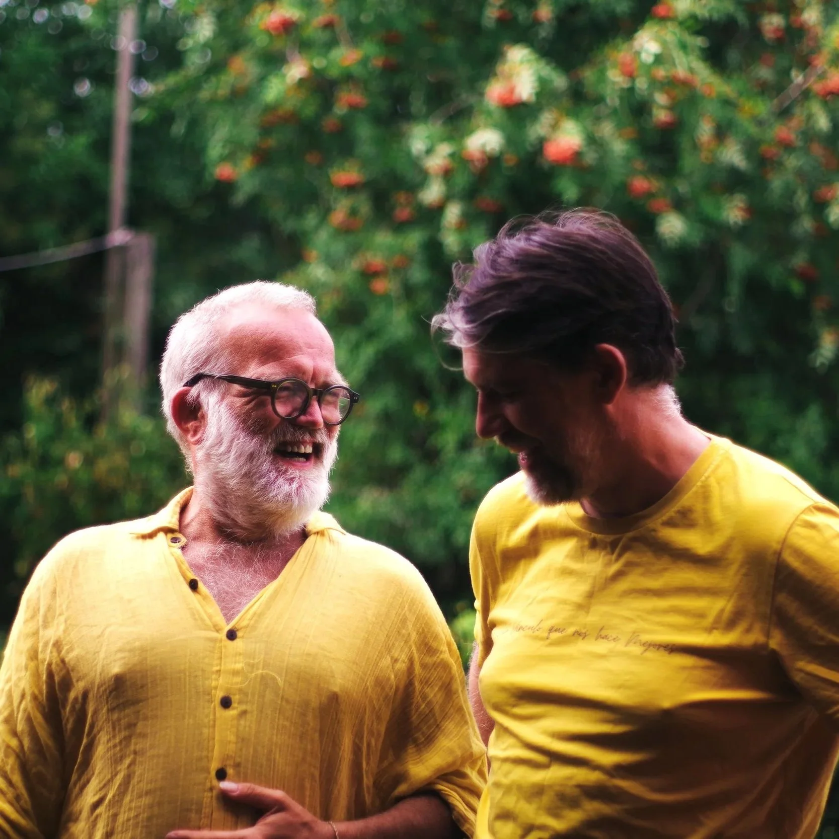 Two men in yellow shirts laughing and smiling outdoors, with a blurred green background of trees and flowers.