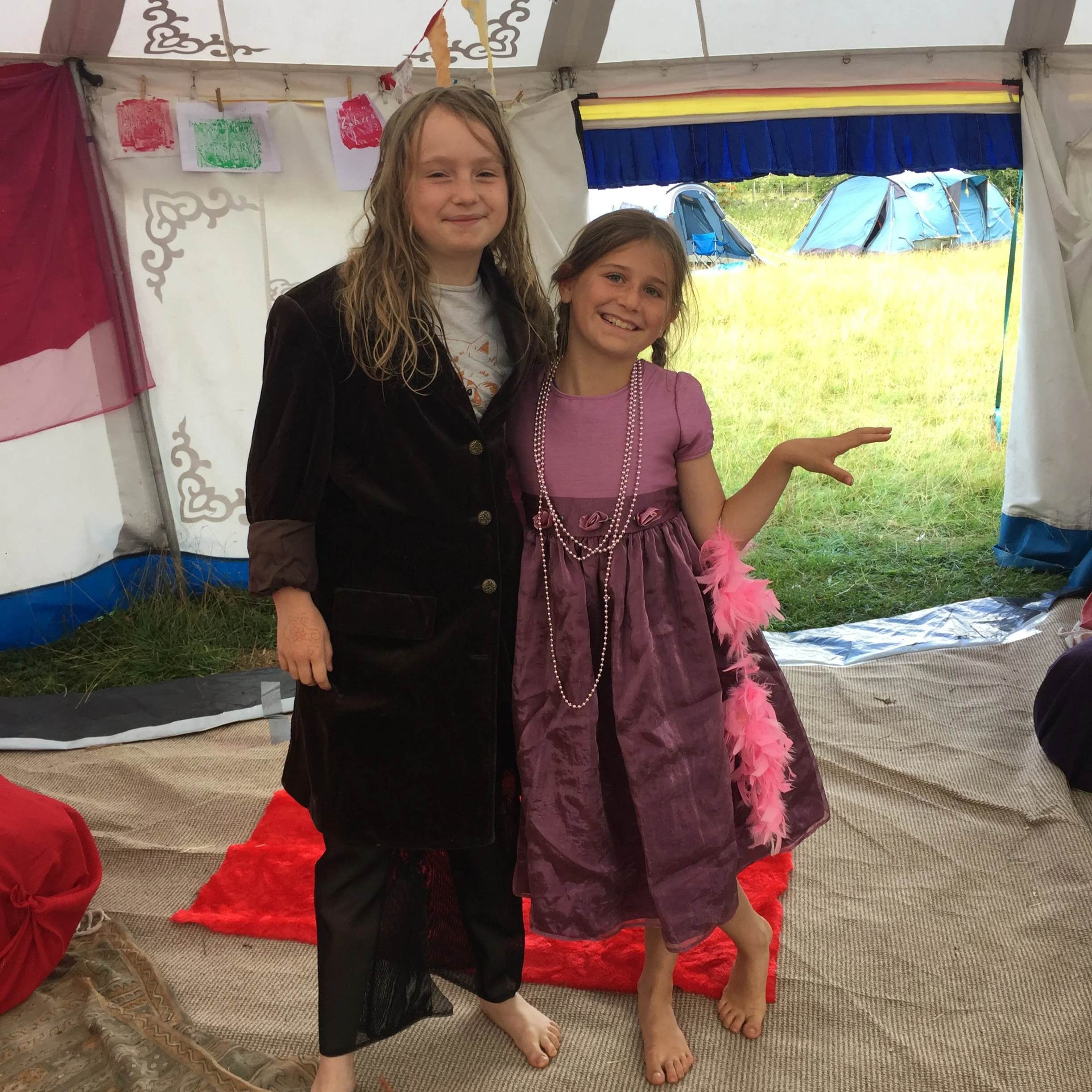 Two young girls standing together indoors, smiling at the camera. One girl has light brown, curly hair and wears a white t-shirt with a black jacket. The other girl has darker hair in braids, a pink dress, and wears pearl necklaces and pink feathered accessories. They are barefoot on a carpeted floor, and a tent with colorful decorations and tents in a grassy outdoor setting are visible in the background.