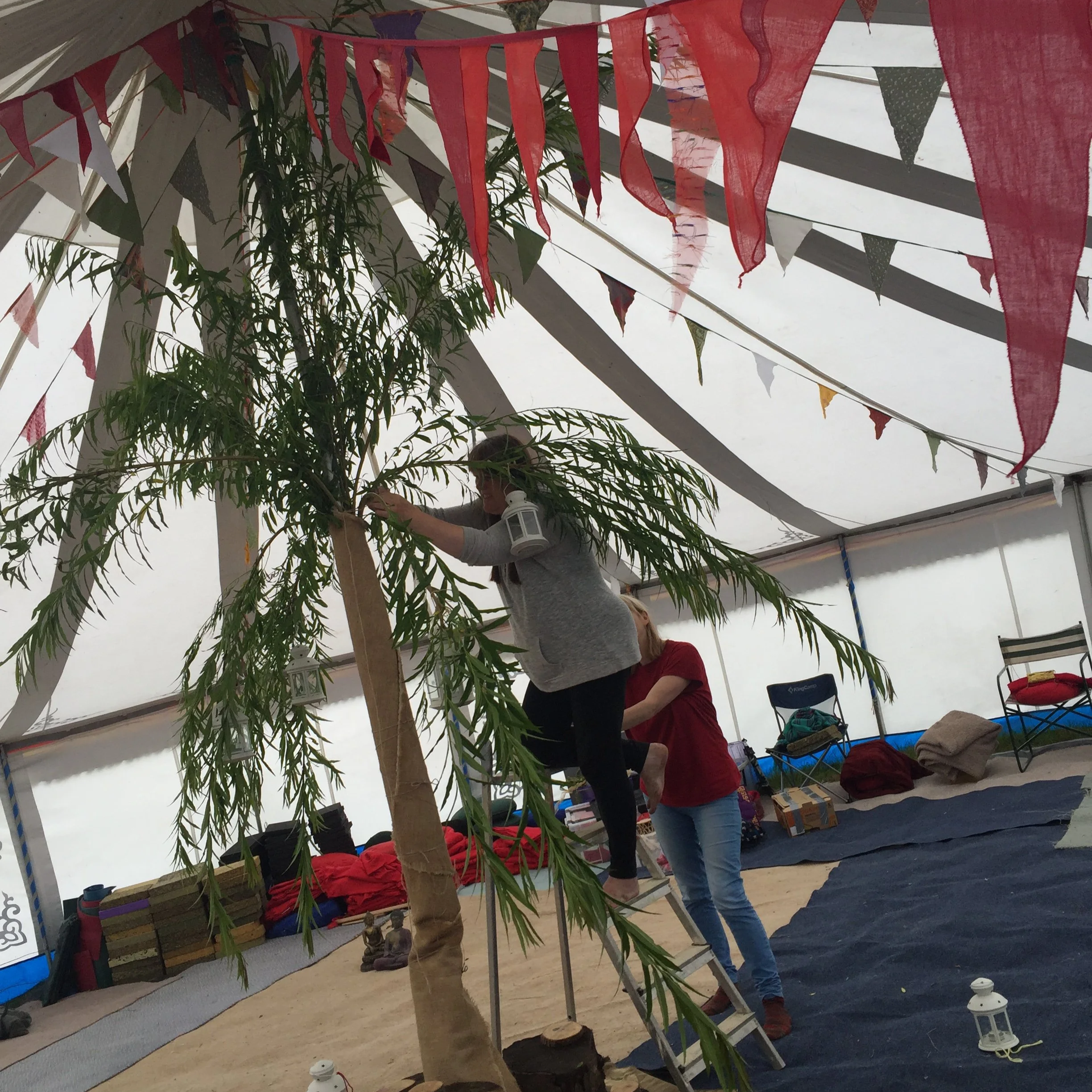 Festive indoor event with colorful bunting banners hanging under a tent-like roof, green plant in the foreground, and people standing and sitting around with chairs and boxes on the floor.