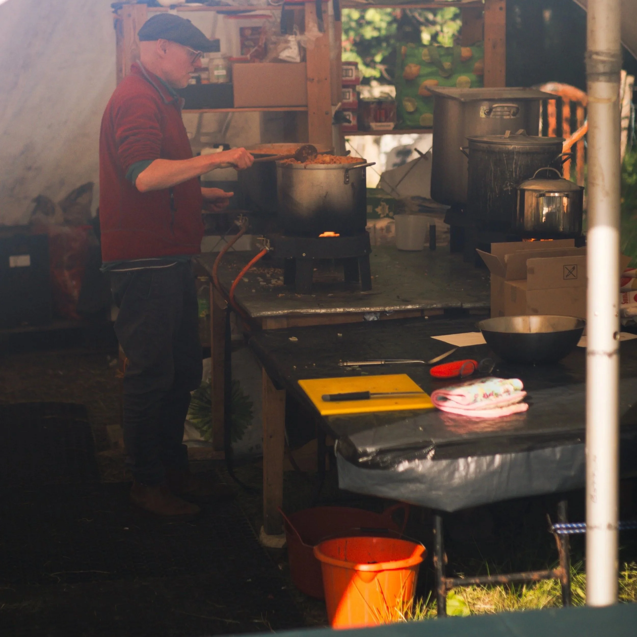 A person wearing glasses and a red jacket is cooking food in a large silver pot at an outdoor setting during daylight. There are boxes and a cloth on a table nearby, with a background of green trees and a structure made of wooden beams.