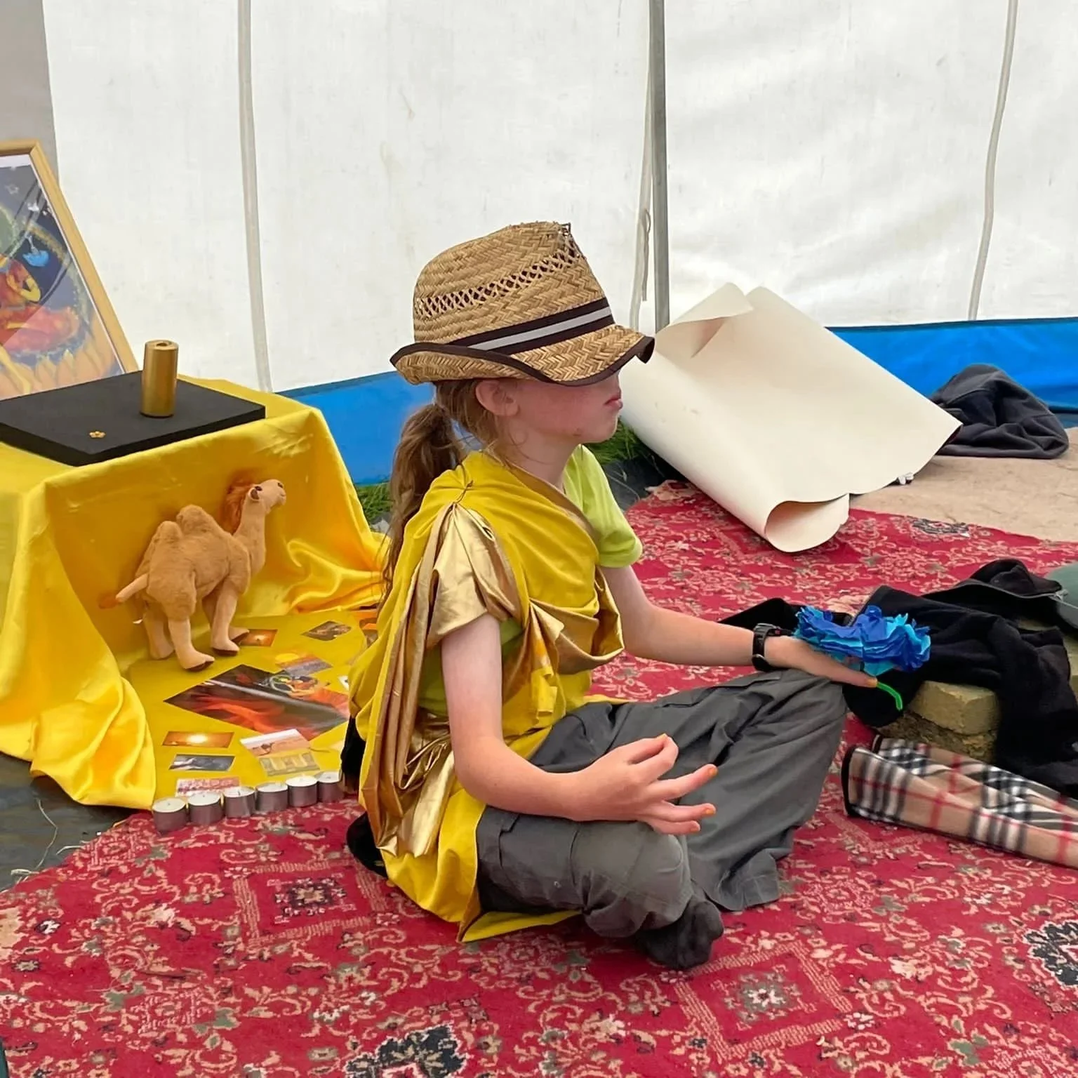 A girl wearing a straw hat and a golden shiny dress, sitting cross-legged on a colorful red carpet inside a tent. She is holding her lap with one hand and appears to be participating in an activity. Behind her, there is a yellow table with a stuffed camel toy, photographs, a framed picture, and various items. The tent's interior shows a white wall and some cloth or paper rolled up, with a bag and other belongings nearby.