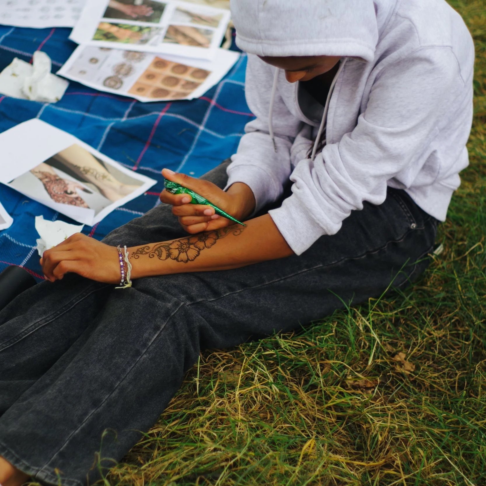 A person sitting on grass, getting a tattoo on their forearm with black floral designs, while there are various papers with images and instructions on a picnic blanket nearby.