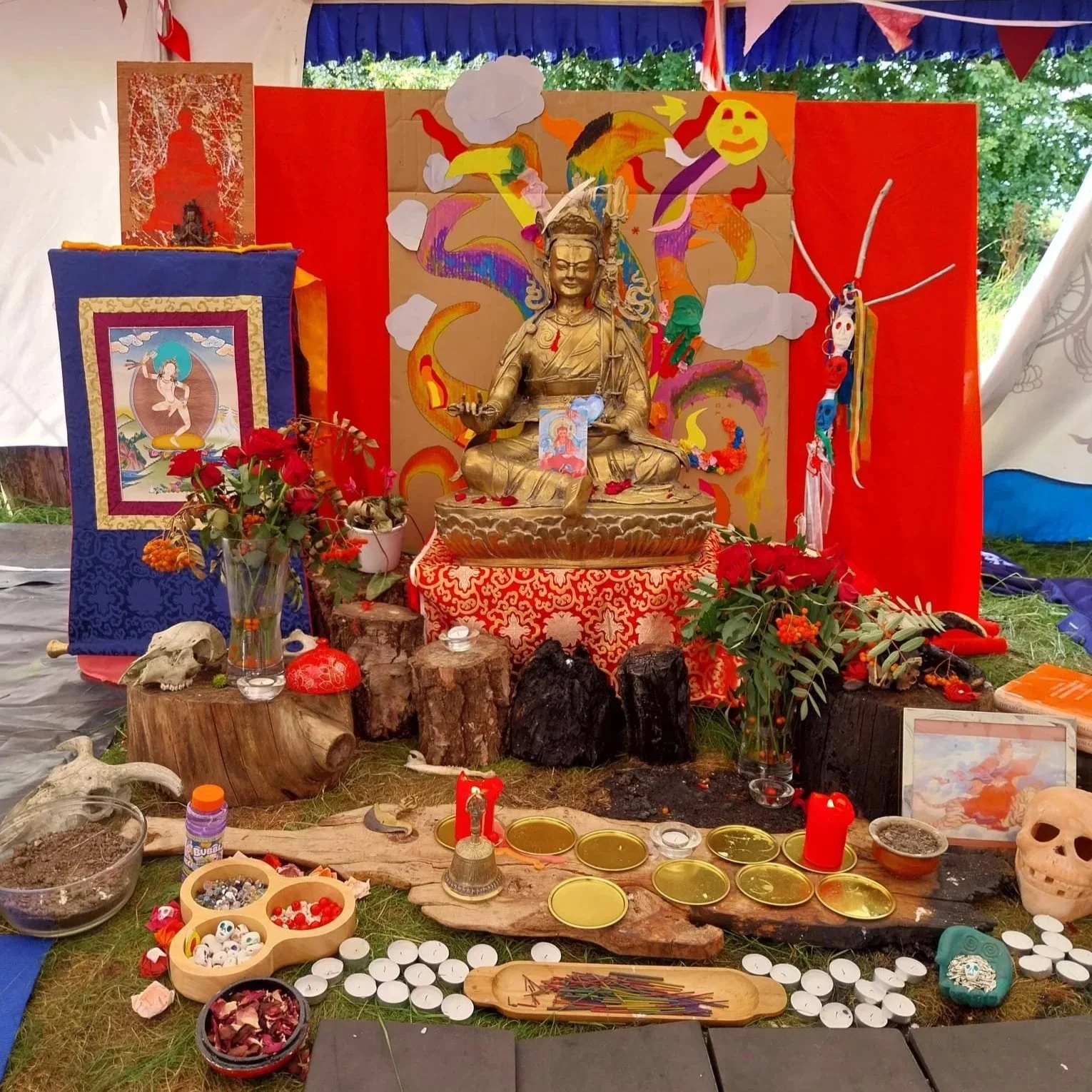 An altar with a golden statue of a deity, decorated with colorful banners, flowers, and candles. Surrounding objects include skulls, bowls with offerings, and painted images, all set outdoors with trees visible in the background.