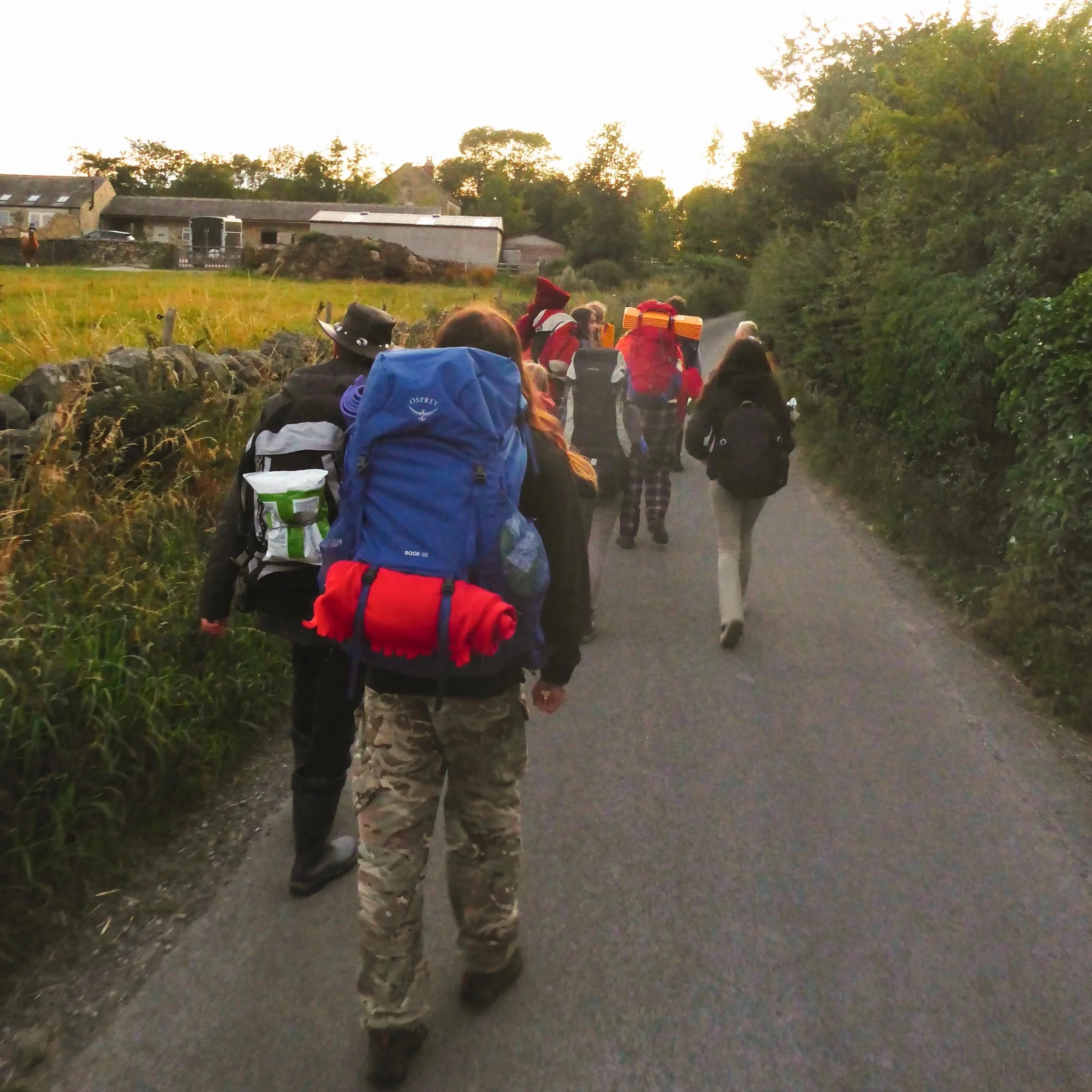 A group of people walking on a rural road during sunset, carrying backpacks and outdoor gear.