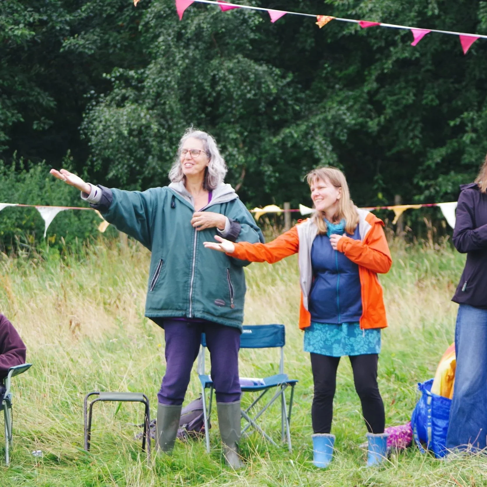 Two women standing outdoors in a grassy field decorated with pink and yellow bunting. One woman is gesturing with her right arm while the other woman is smiling and looking at her. They are dressed in casual weather-appropriate clothing.