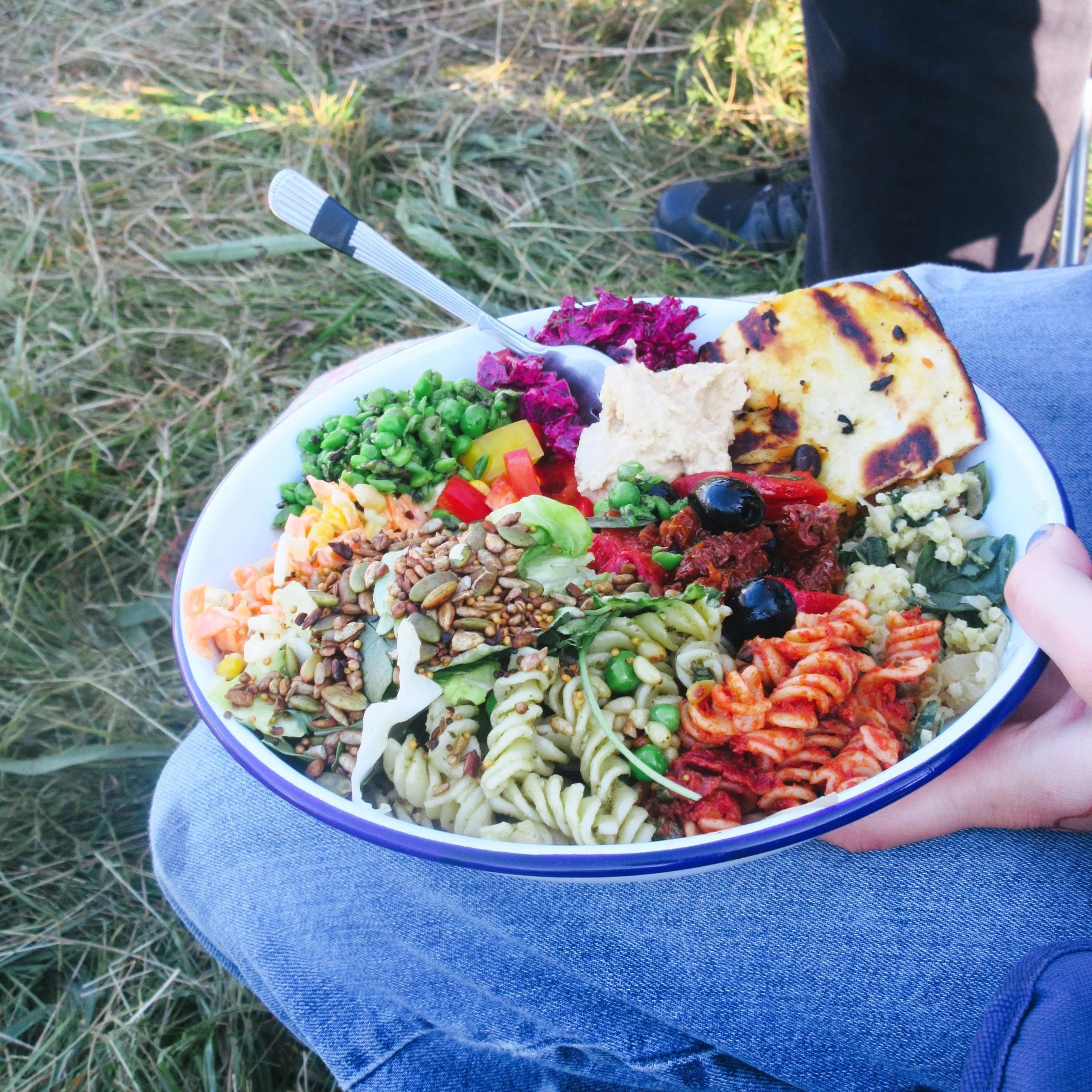 A plate of assorted salads and grilled vegetables being held over grass and dirt, with a person wearing black shoes and jeans visible in the background.