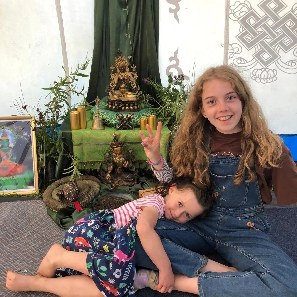 Two young girls, one smiling and posing with a peace sign, sitting on the floor in front of a display of Buddhist statues and artifacts. The older girl has long, curly blonde hair and is wearing denim overalls, while the younger girl has straight hair and is wearing a patterned dress. The display includes seated and standing figurines, candles, plants, a snake, and a framed picture of a deity.