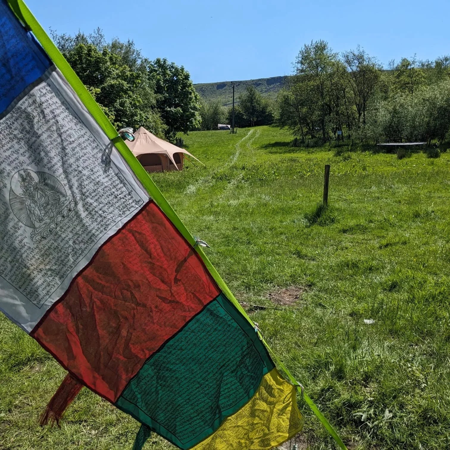 Colorful prayer flag with intricate text and images, set against a lush green outdoor landscape with trees, a tent, and a mountain in the background under a blue sky.